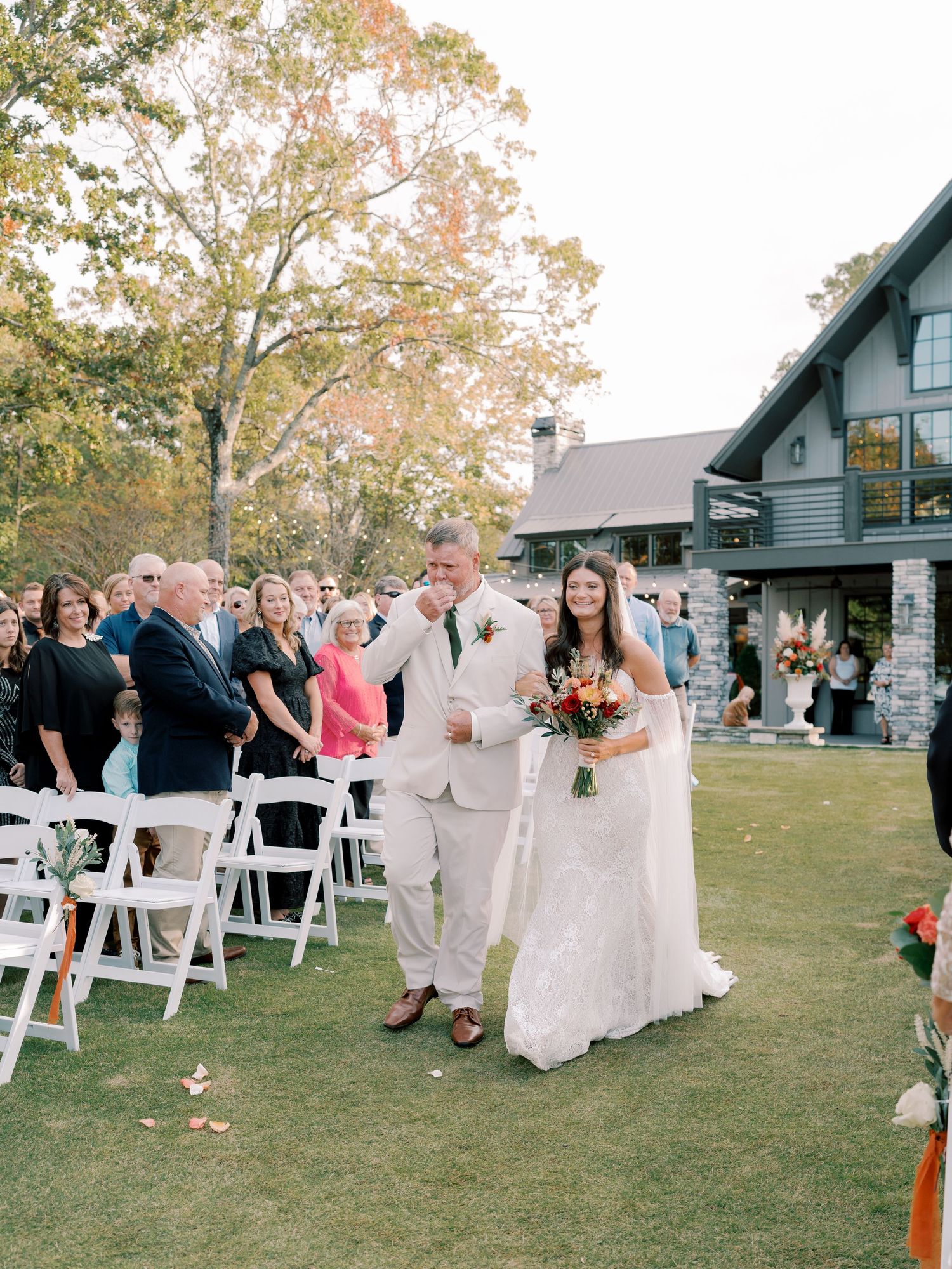 Outdoor wedding ceremony setup with white chairs and guests on a lawn.