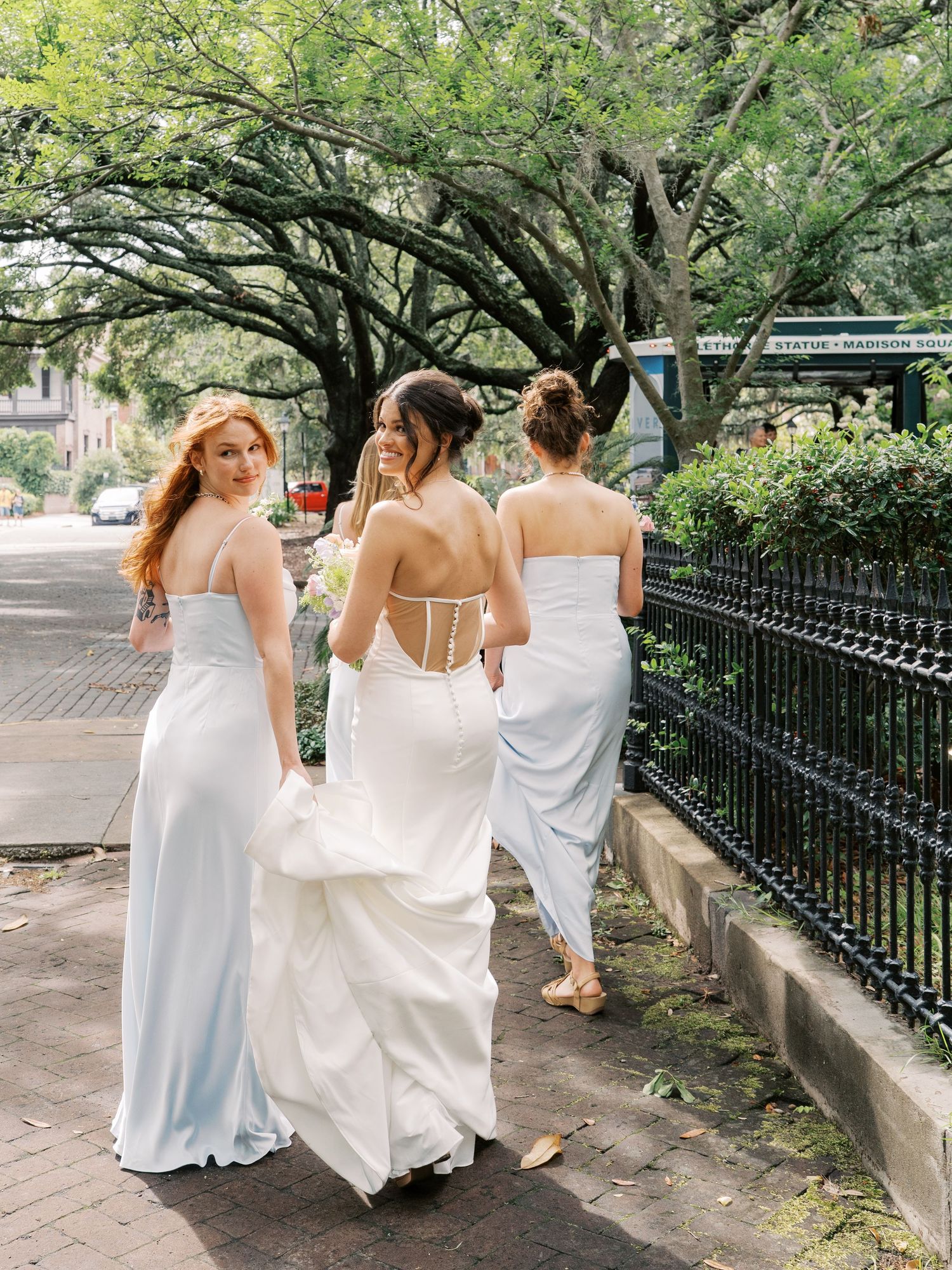 Bridesmaids in white dresses walk together on a tree-lined path with iron fencing.