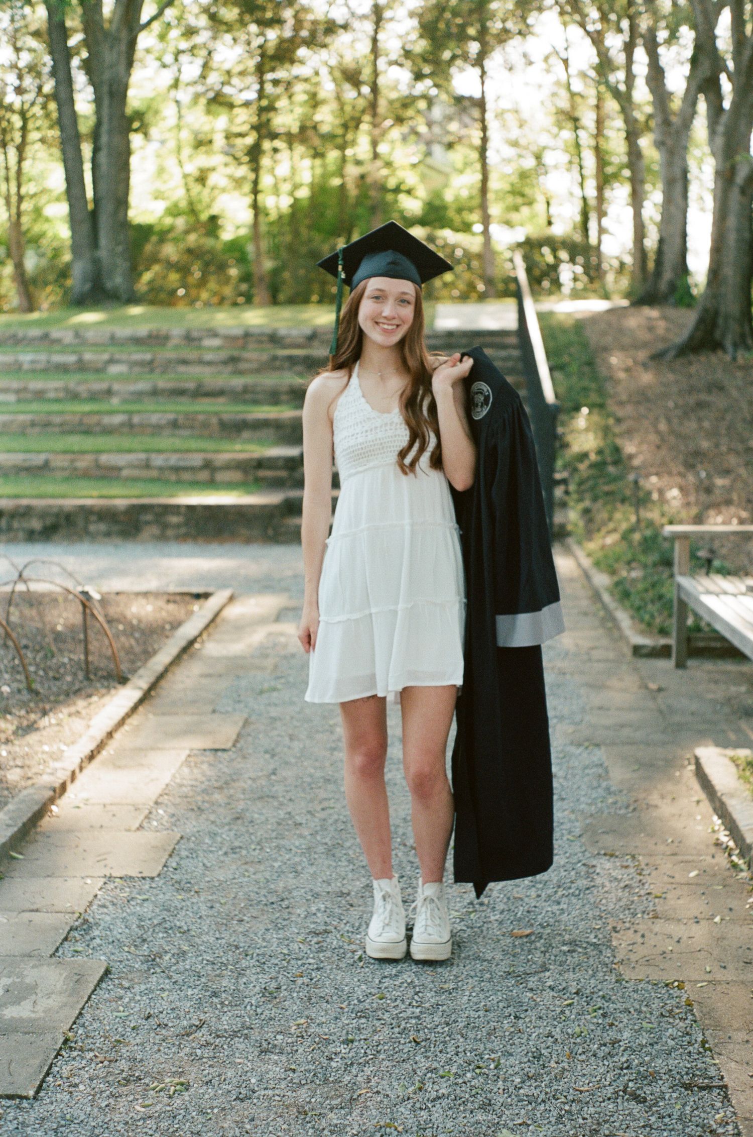 Graduate in white dress and cap holding graduation gown on stone garden steps.