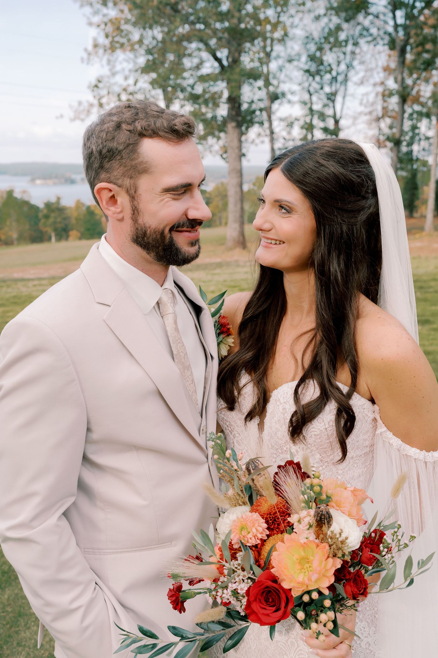 Newlyweds share a romantic moment outdoors with autumn foliage and a vibrant red and peach bouquet.