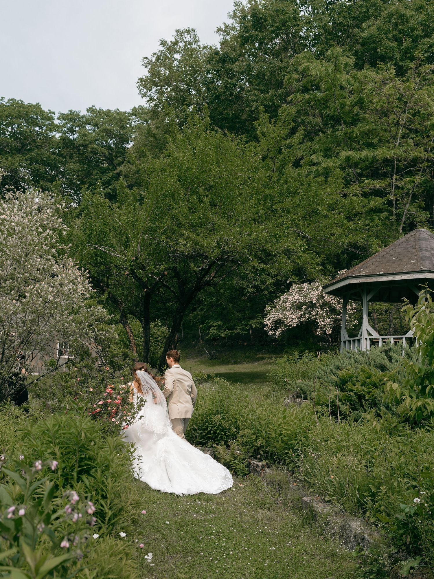 A wedding couple walks through a garden path with flowering trees and a gazebo in the background.