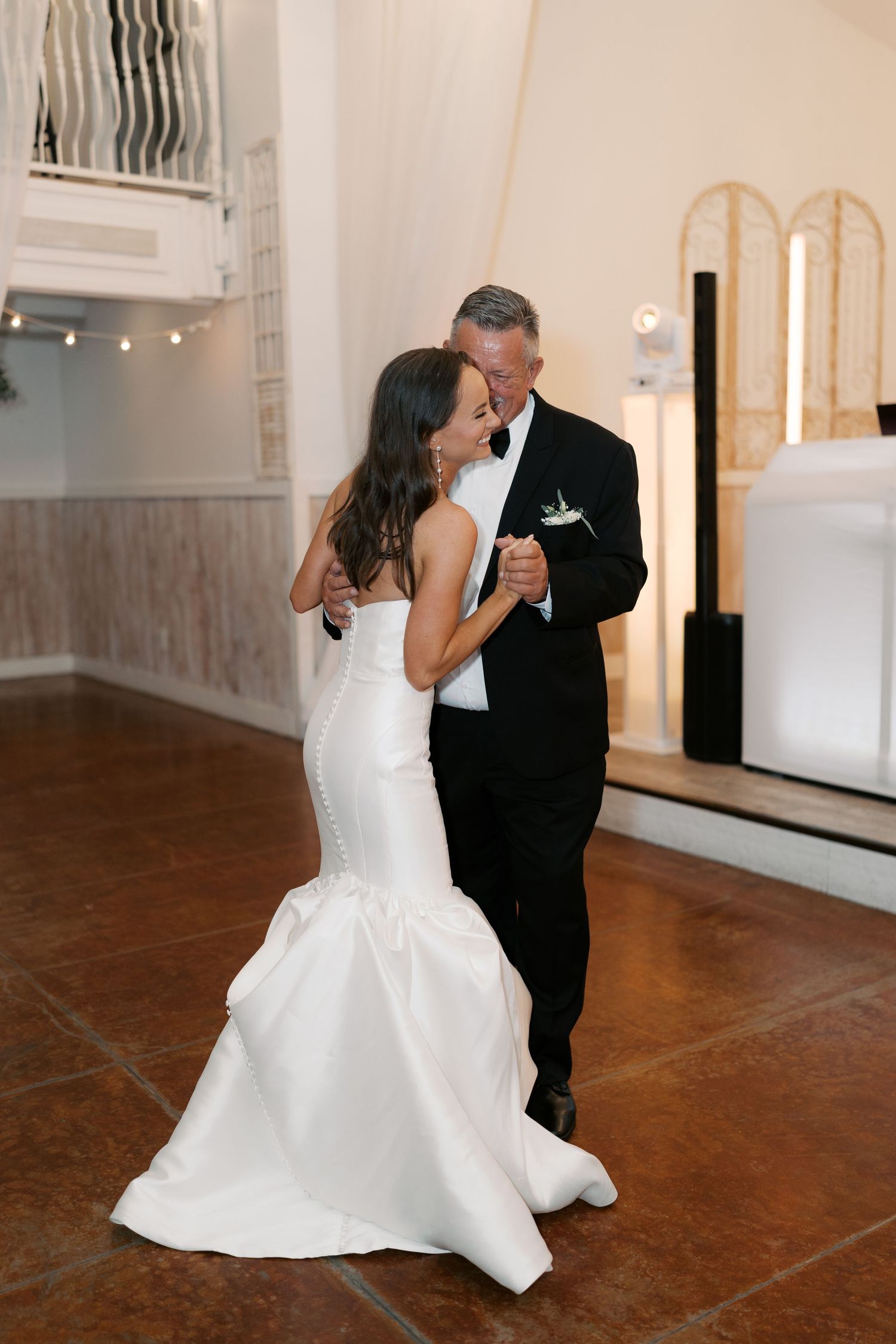A couple shares an intimate dance moment in an elegant white venue with hardwood floors.