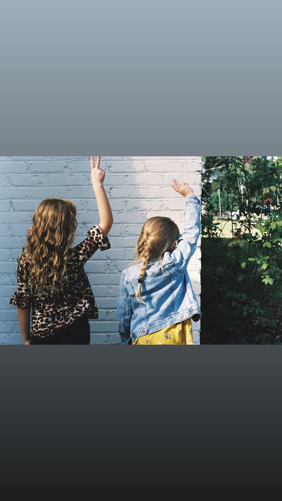 Two friends raising their hands joyfully against a white brick wall on a sunny day.
