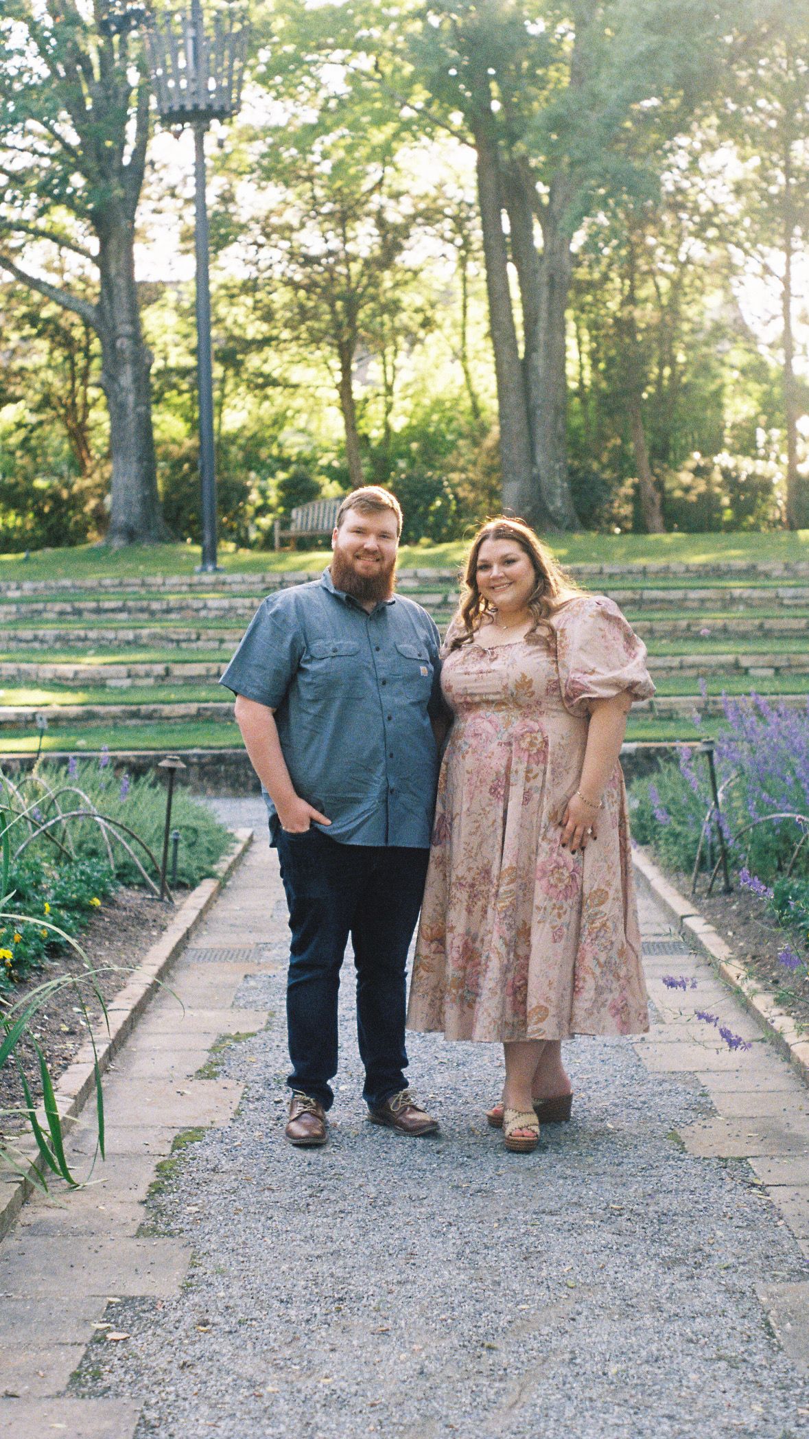 Couple poses together on a gravel path surrounded by greenery in an outdoor garden setting.