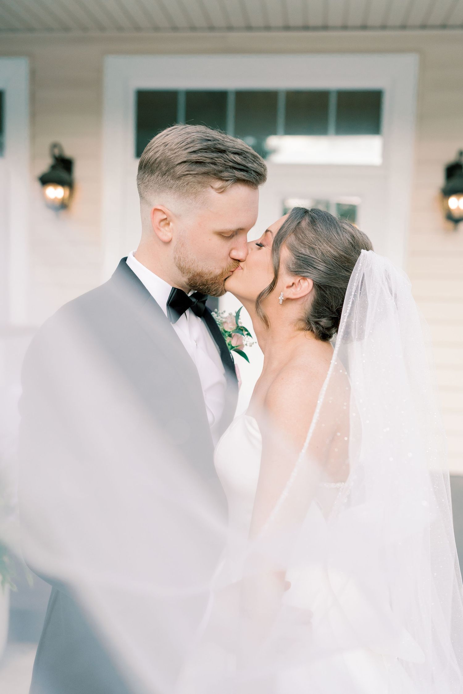 Romantic wedding couple shares an intimate kiss in front of a white building with lanterns.