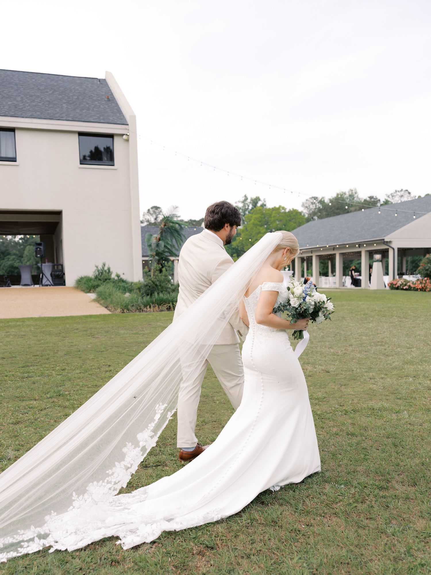 A flowing cathedral veil trails behind a bride in a white dress on a grassy field.
