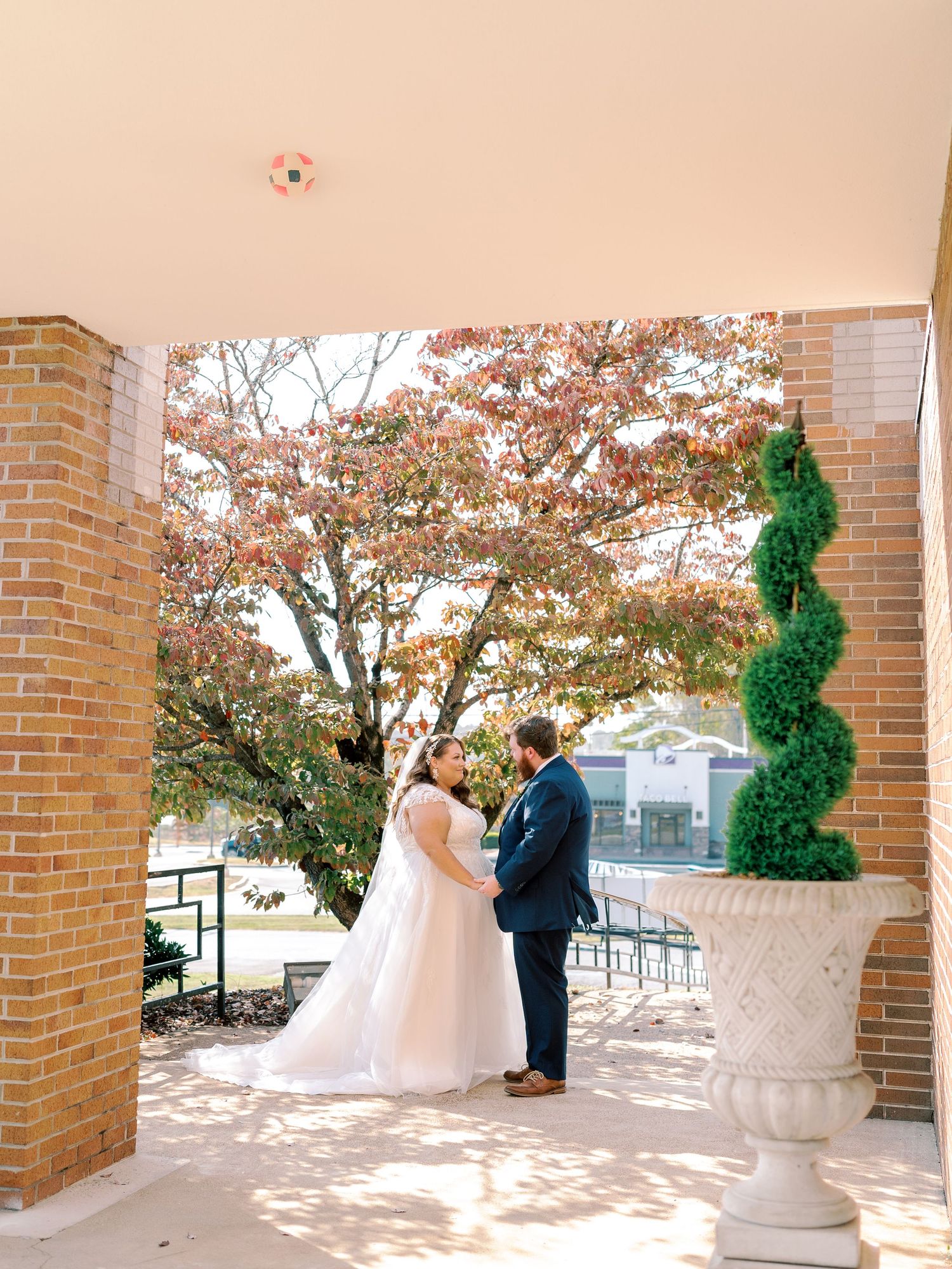 A wedding couple stands together on a porch with brick columns and decorative planters.