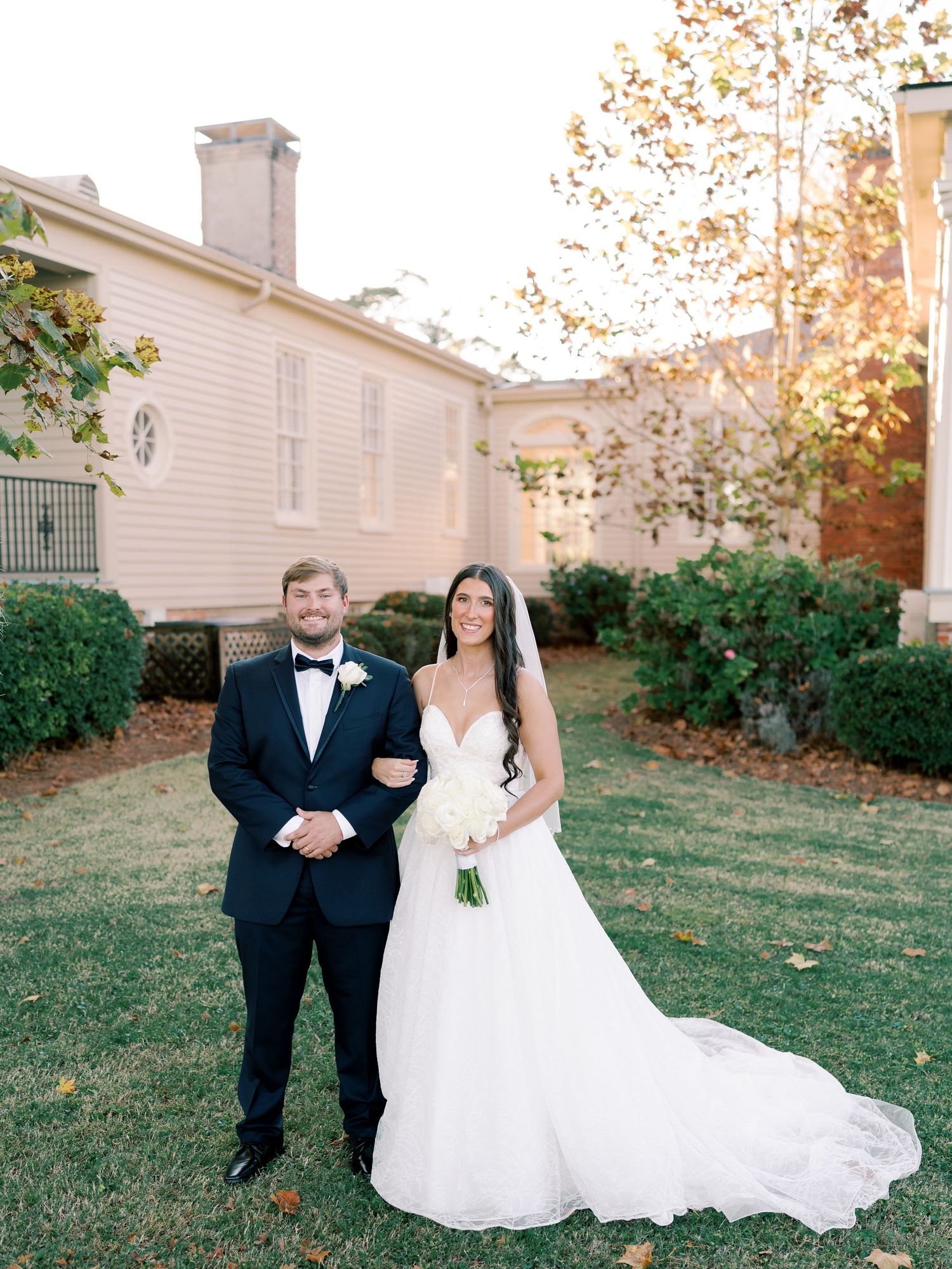 Wedding couple walking together on grass with autumn trees and building in background.