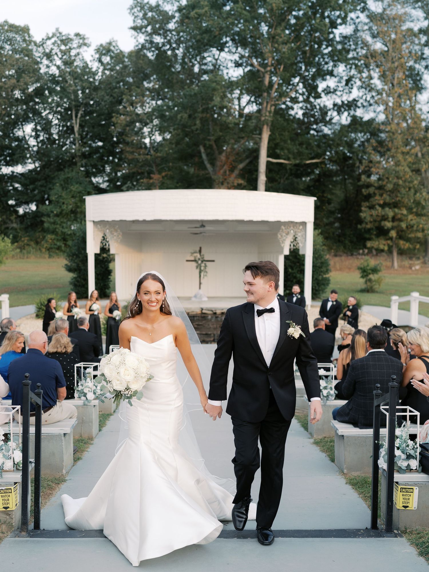 Newlyweds walking down the aisle after ceremony at outdoor chapel with guests seated on either side.