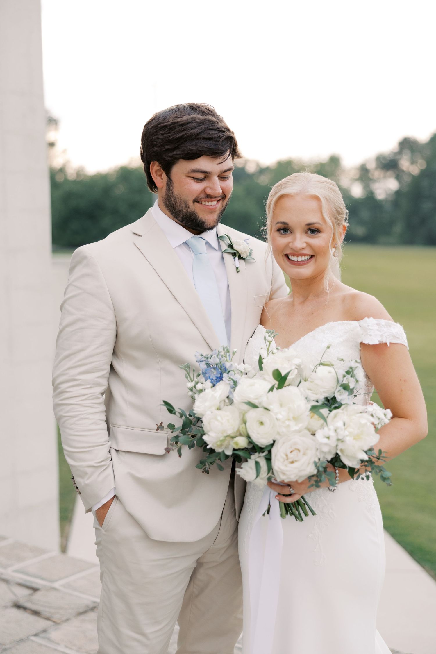 Wedding couple in cream and white attire holding floral bouquets outdoors.