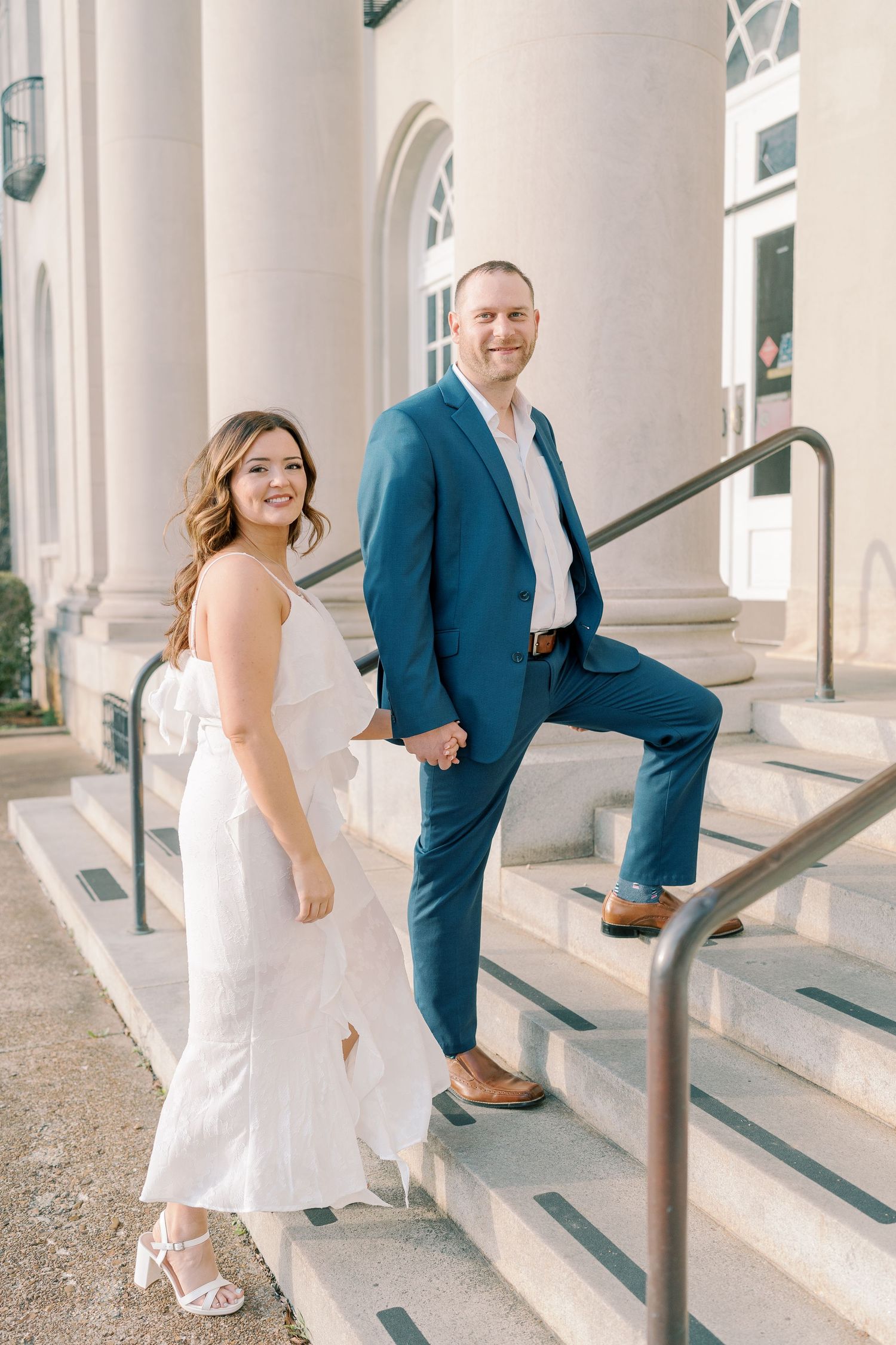 Playful couple poses on white columned porch with steps and handrails.