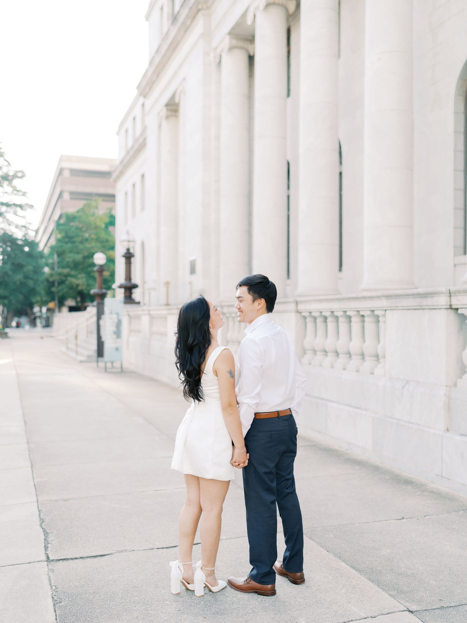 A couple shares playful moments along a classical architecture walkway with white columns.