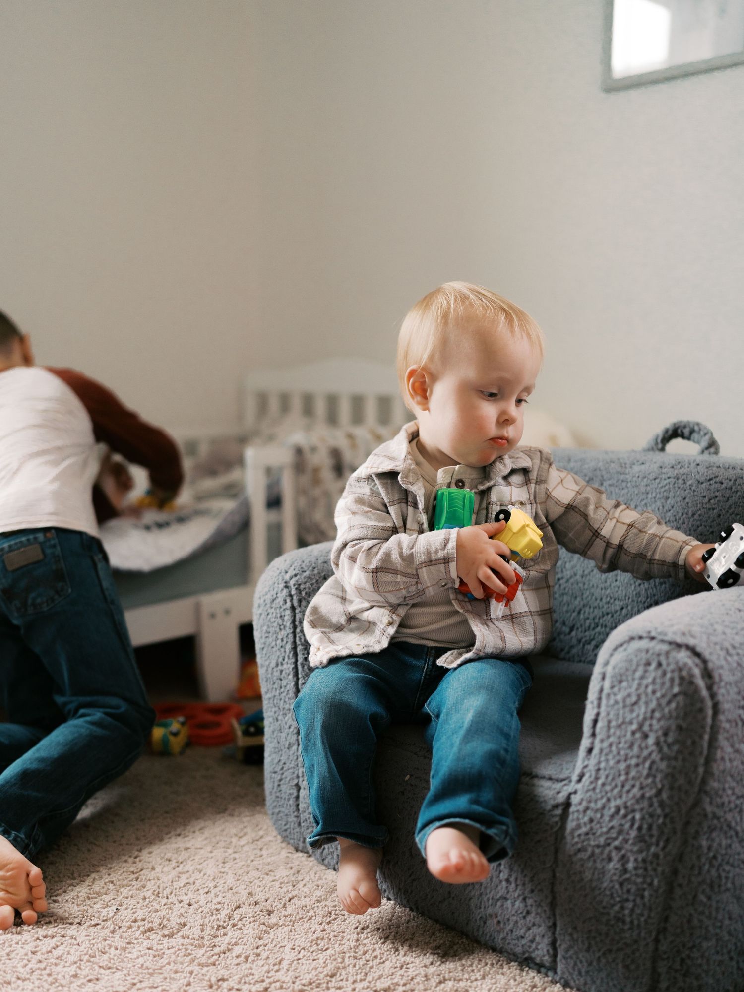 Small child sitting on grey armchair with toys in a cozy nursery setting.