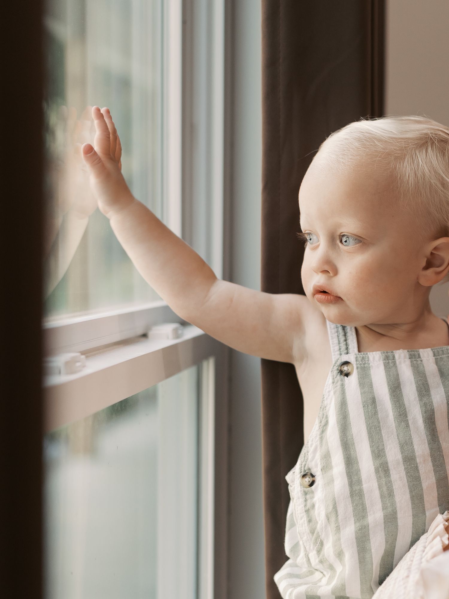 A small child in striped clothing reaches up towards a bright window.
