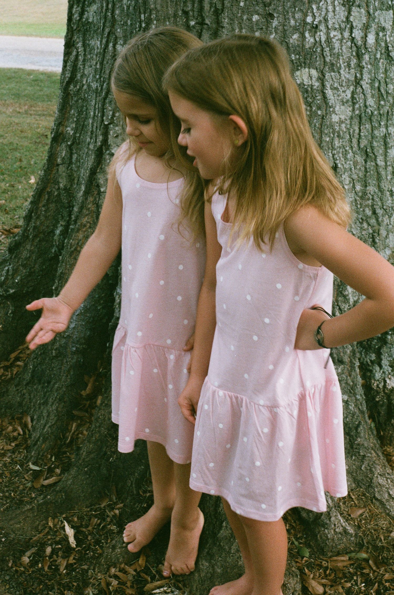 Two children in matching pink polka dot dresses examining something near a tree trunk.