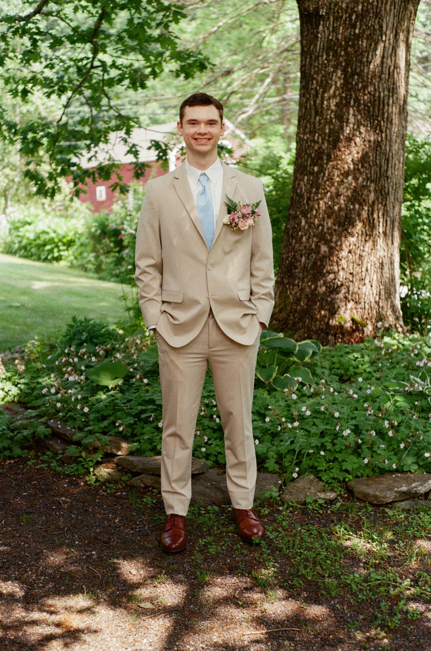 Groomsman in tan suit and blue tie poses under shaded trees in garden setting.