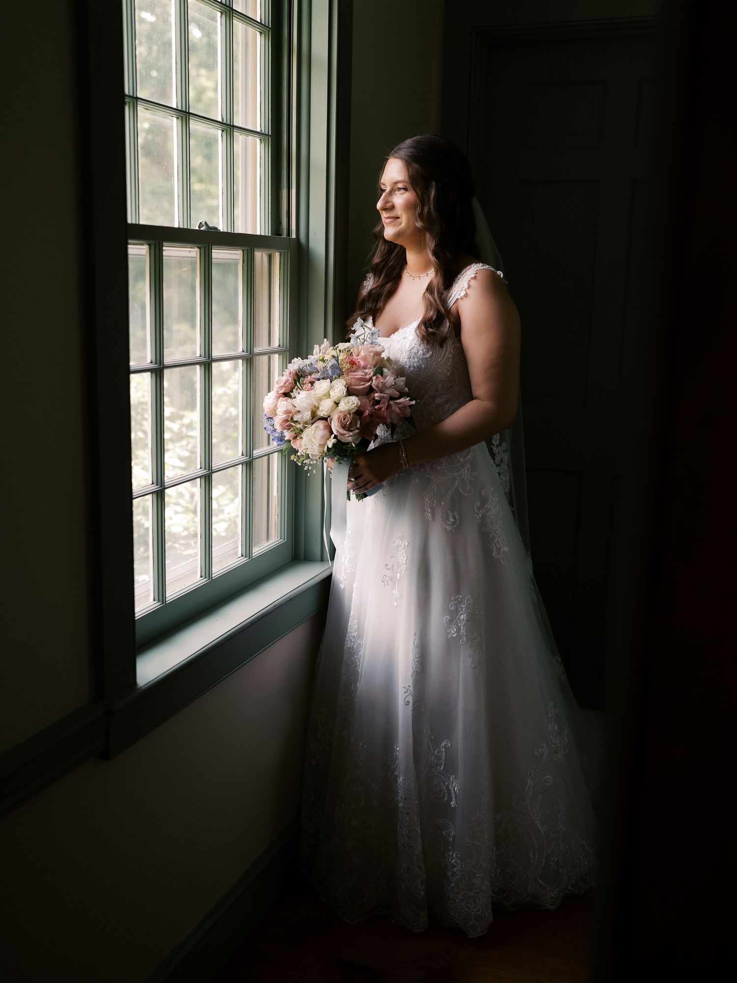 Bride in white gown standing by window holding bouquet in dramatic lighting.