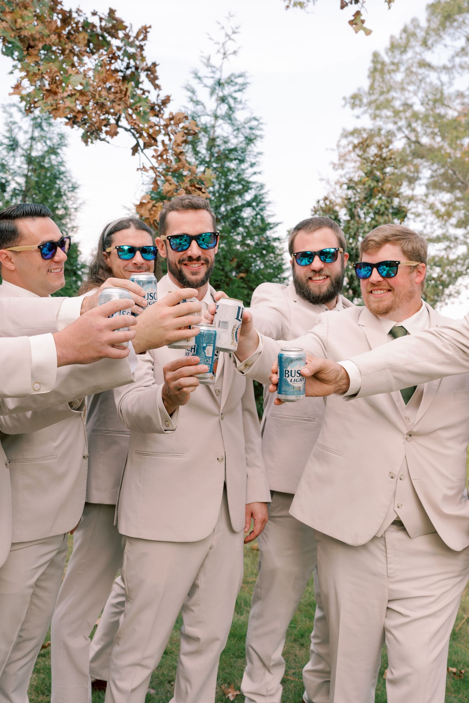 Groomsmen in beige suits wearing sunglasses toasting with drinks outdoors.