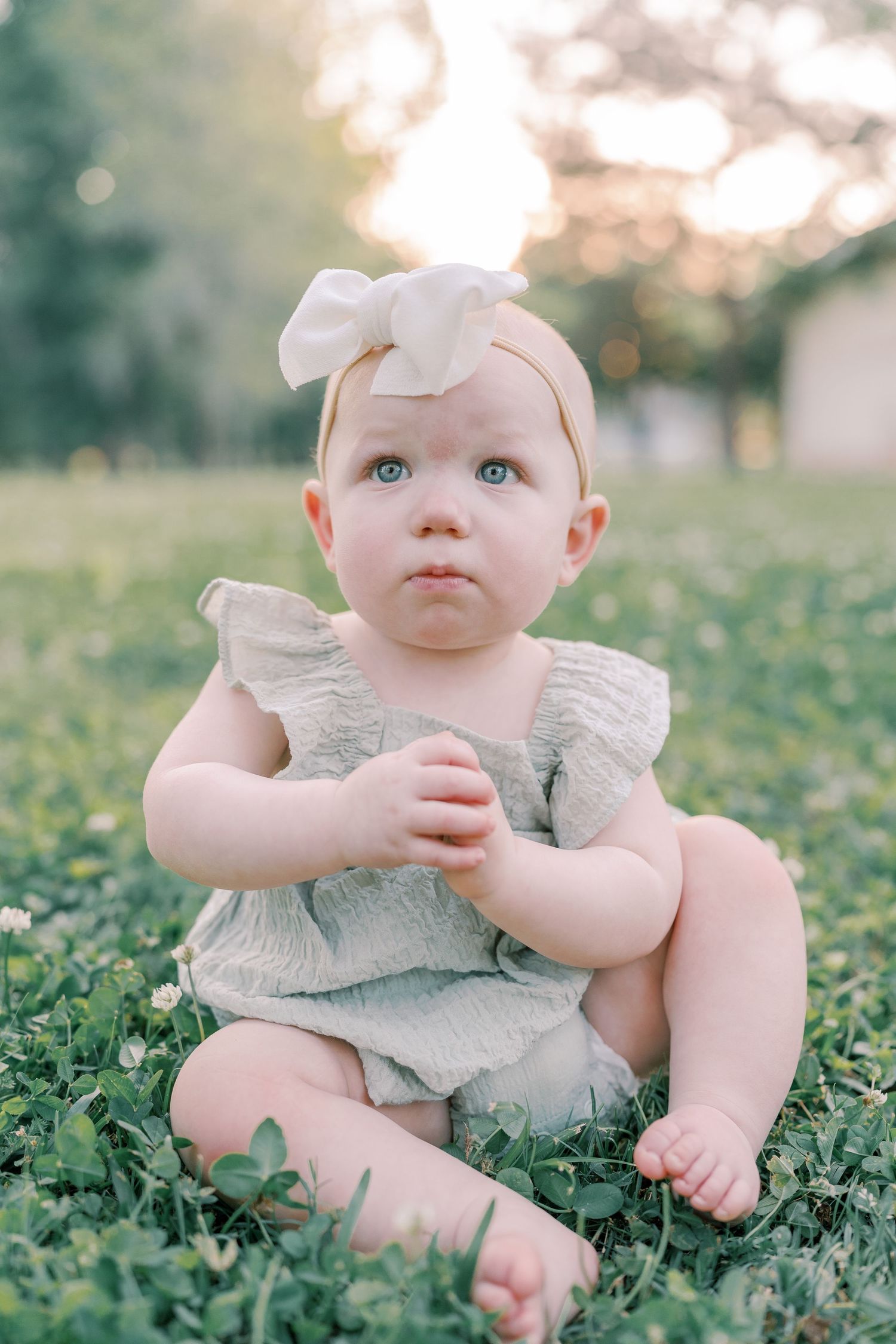 Baby in ruffled dress sits in green grass during golden hour.