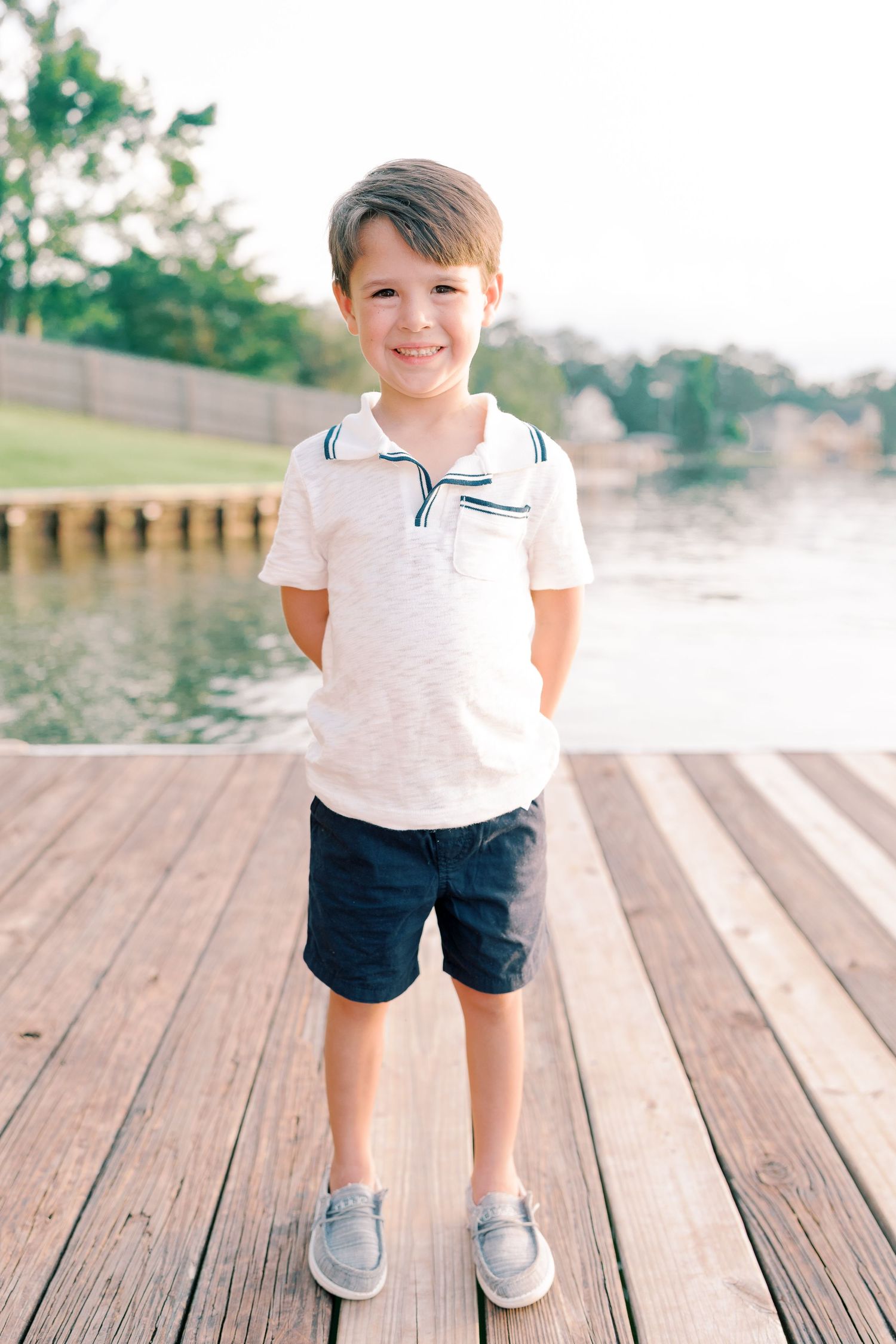Young child in white polo and navy shorts standing on wooden dock.