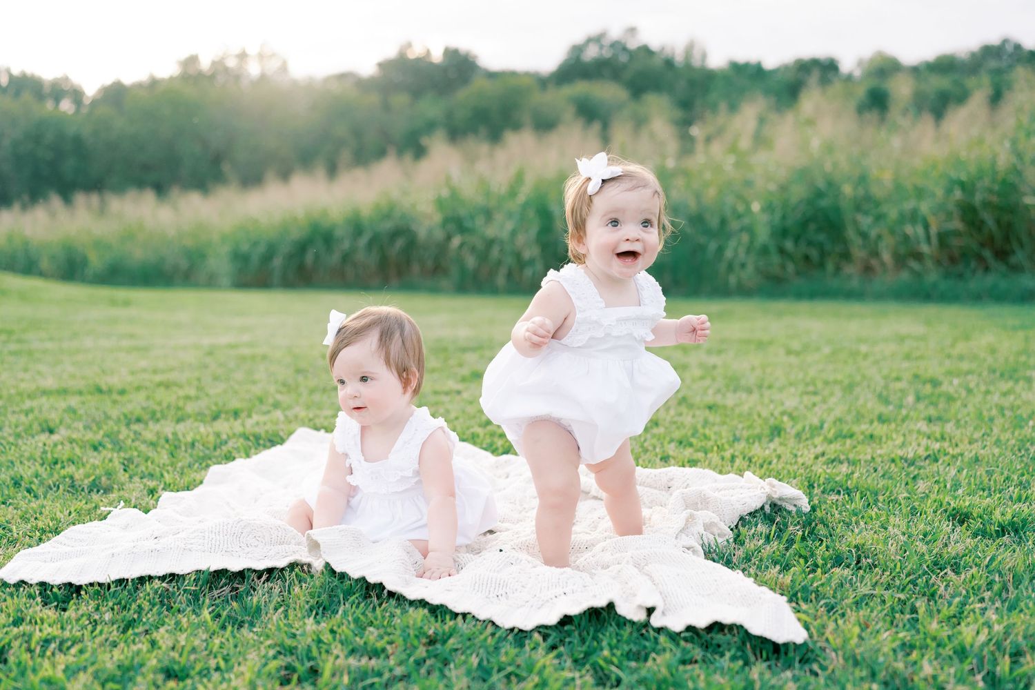 Baby in white dress crawling on blanket in grassy field.