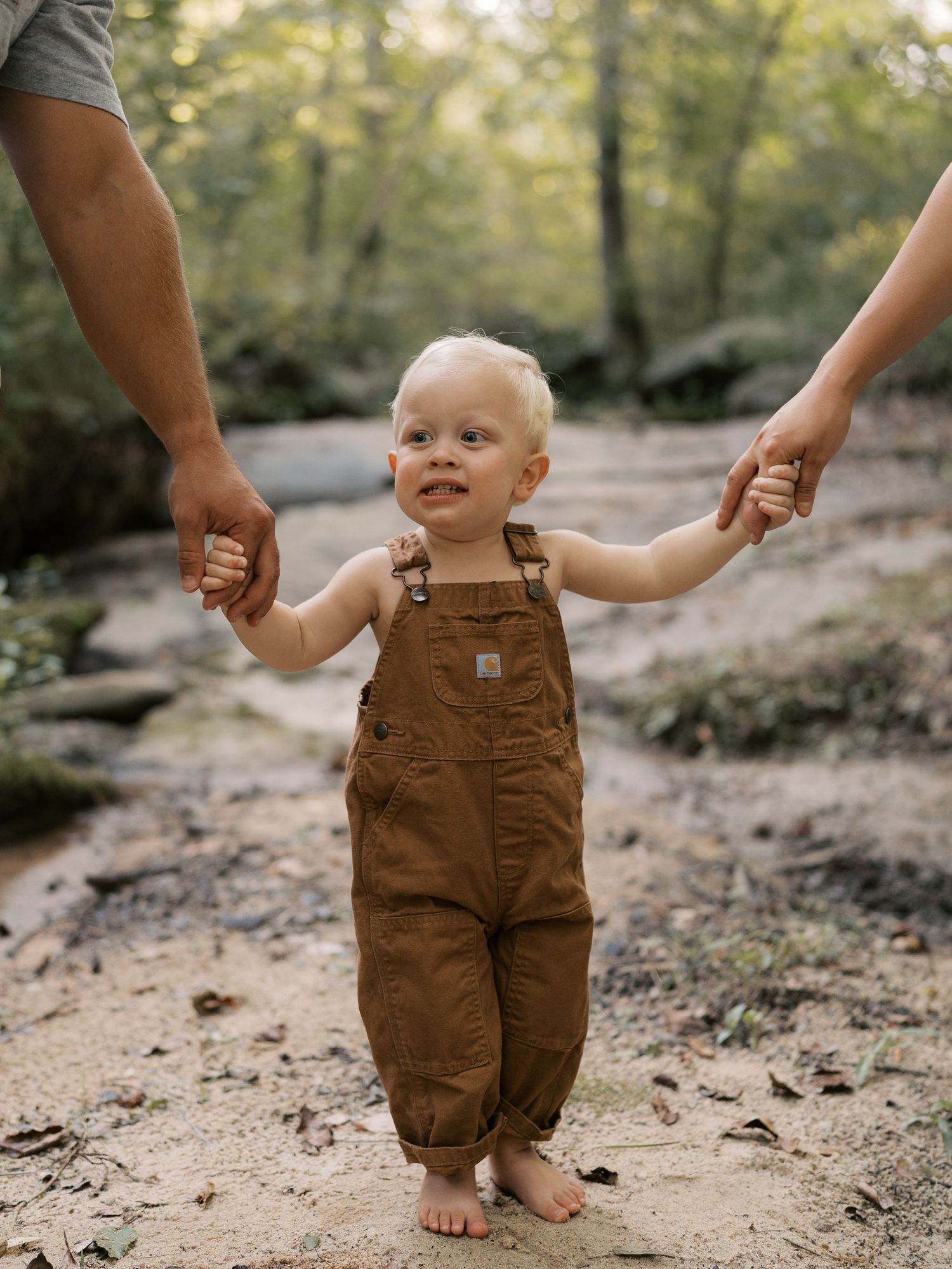 A baby in brown overalls takes first steps on a dirt path in the woods.