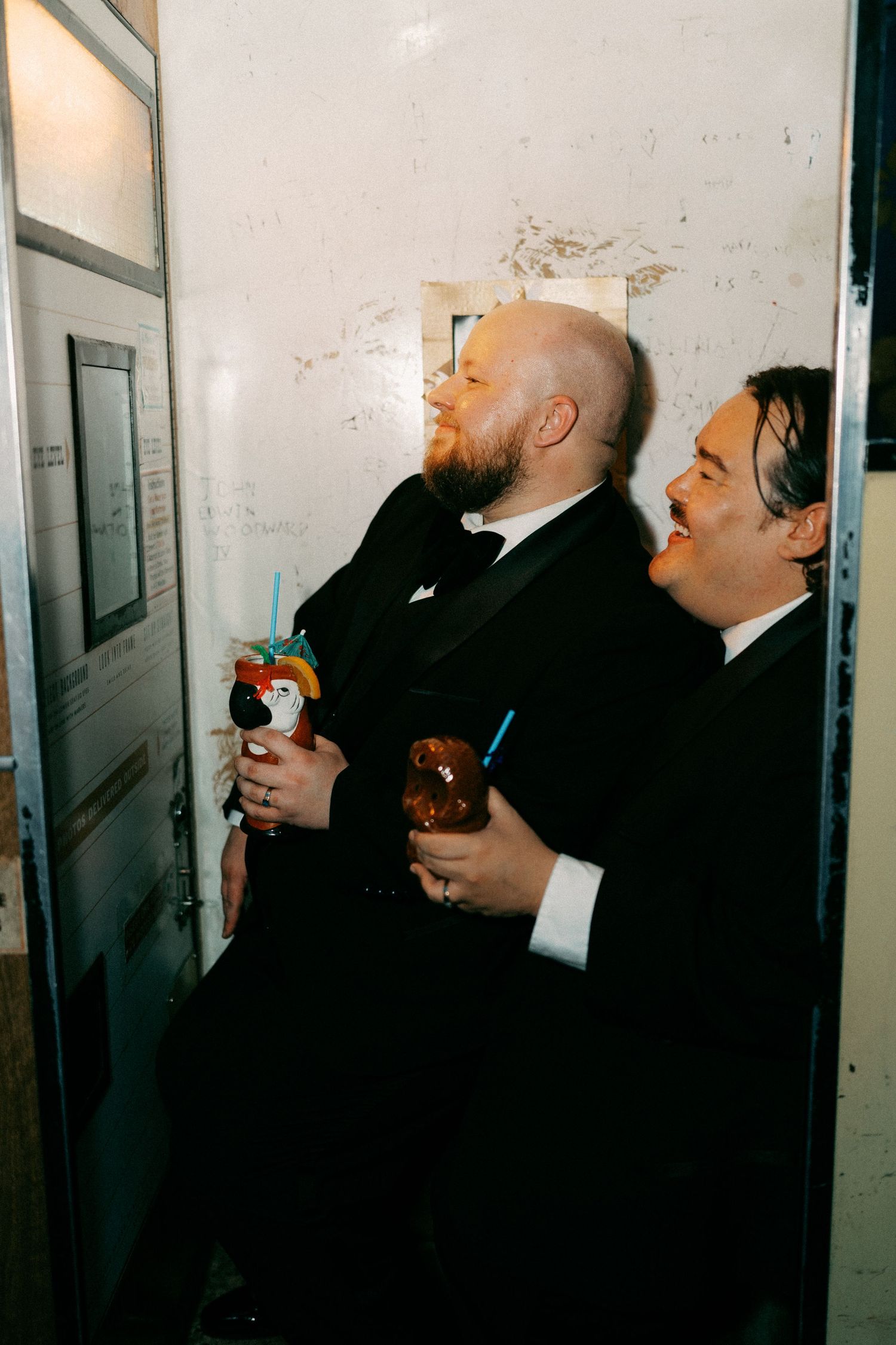 Two people in black suits laughing and enjoying drinks in a hallway.