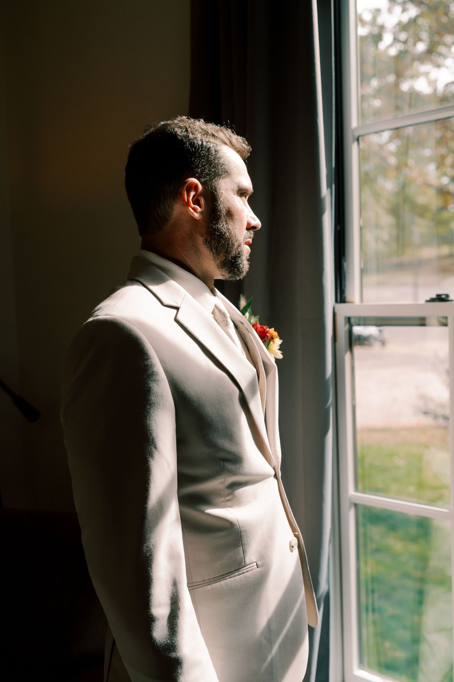 Groom in cream suit looking contemplatively out window in dramatic lighting.