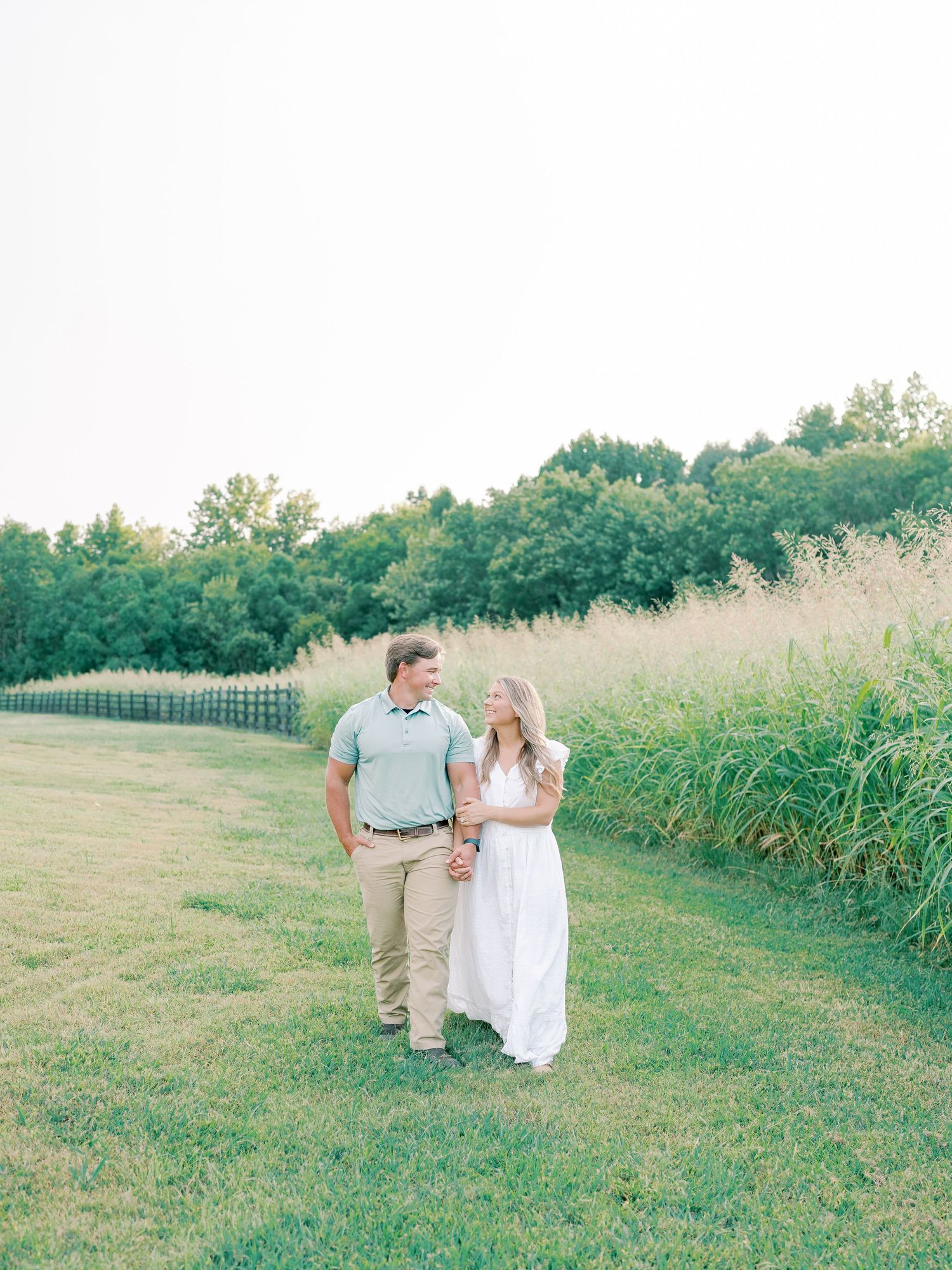 Romantic couple strolling hand in hand through summer field during sunset photo session.