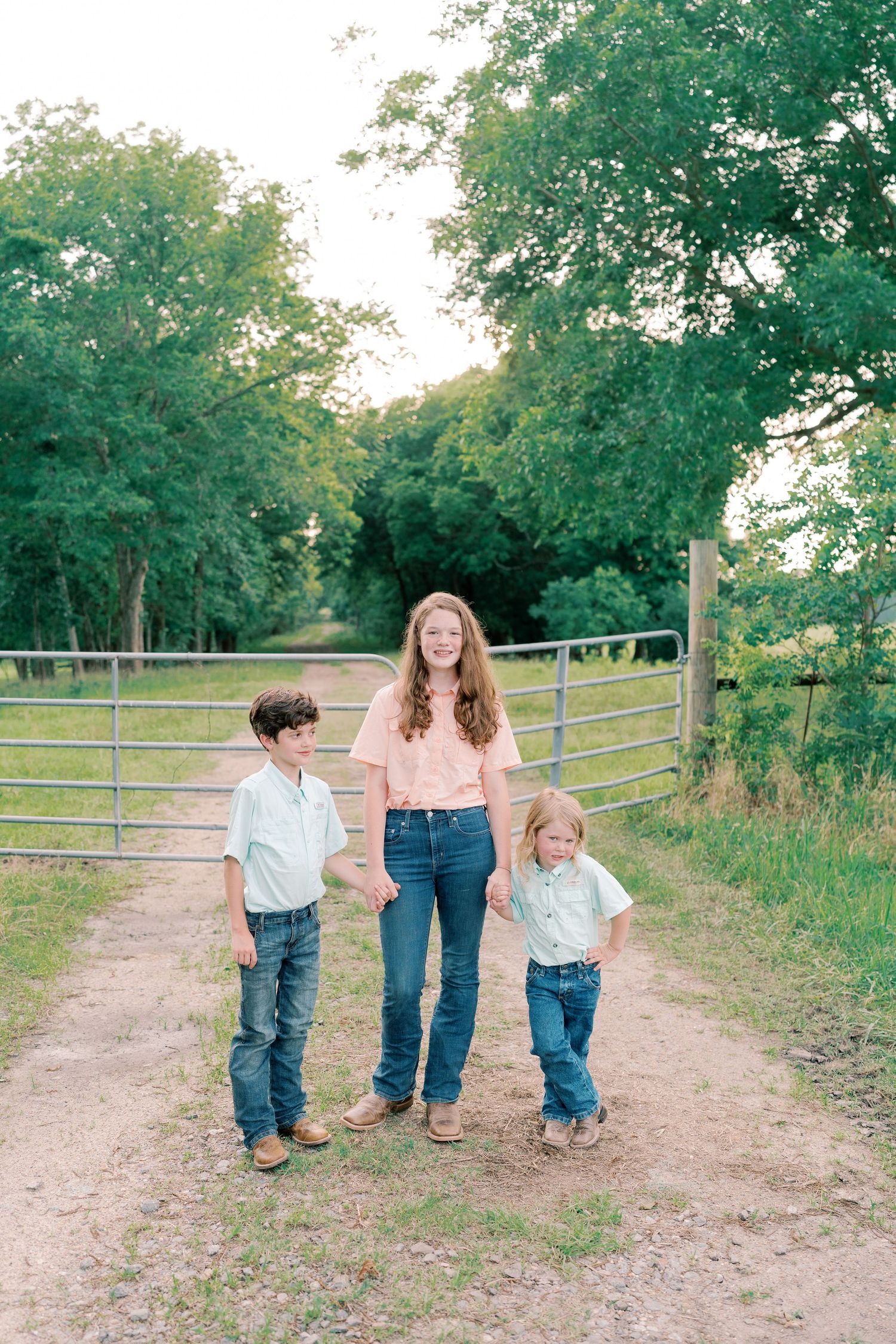 Three people stand together on dirt path with metal gate and trees in background.