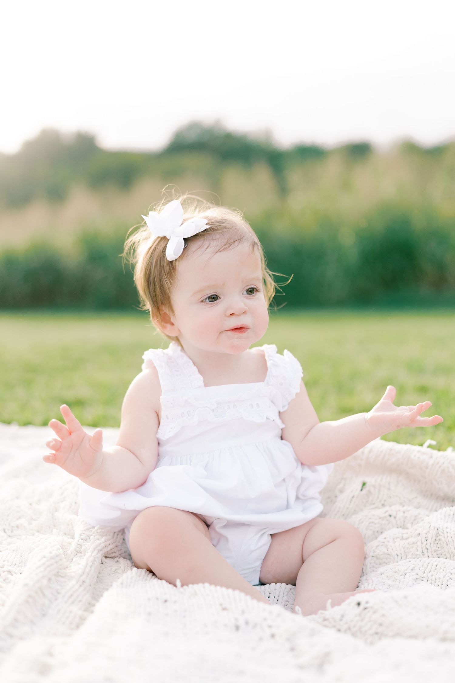 Baby in white dress plays on blanket during outdoor photo session in sunny field.