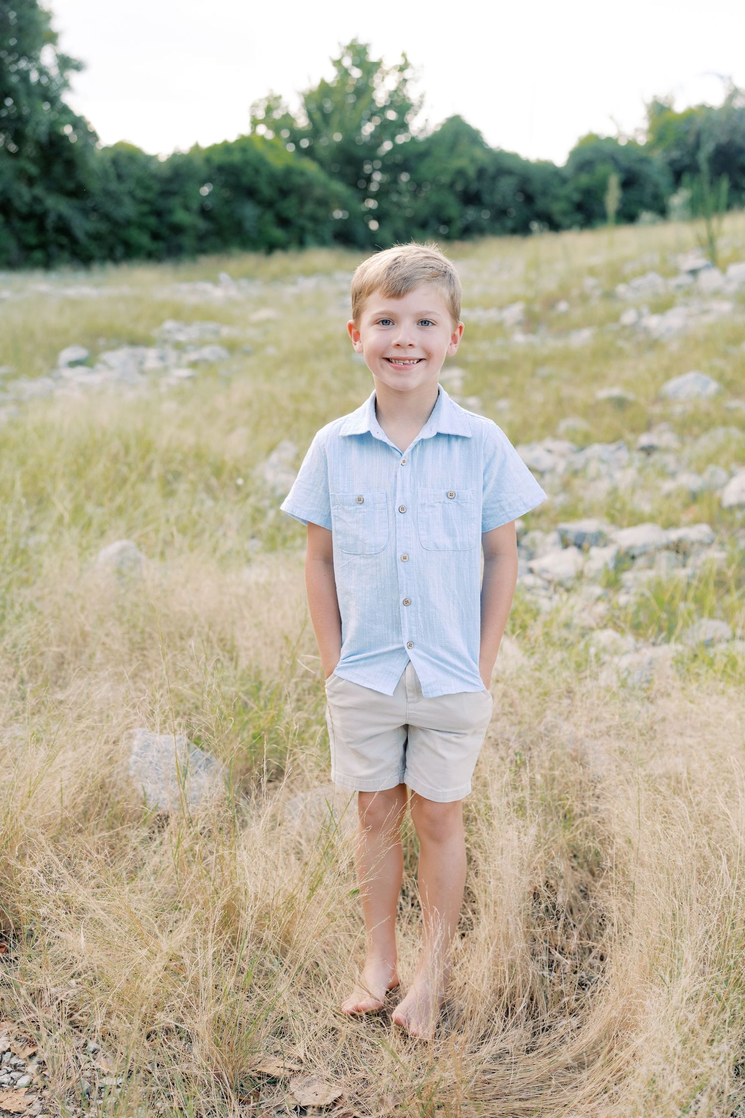 Young boy in light blue shirt and shorts standing in tall grass field.