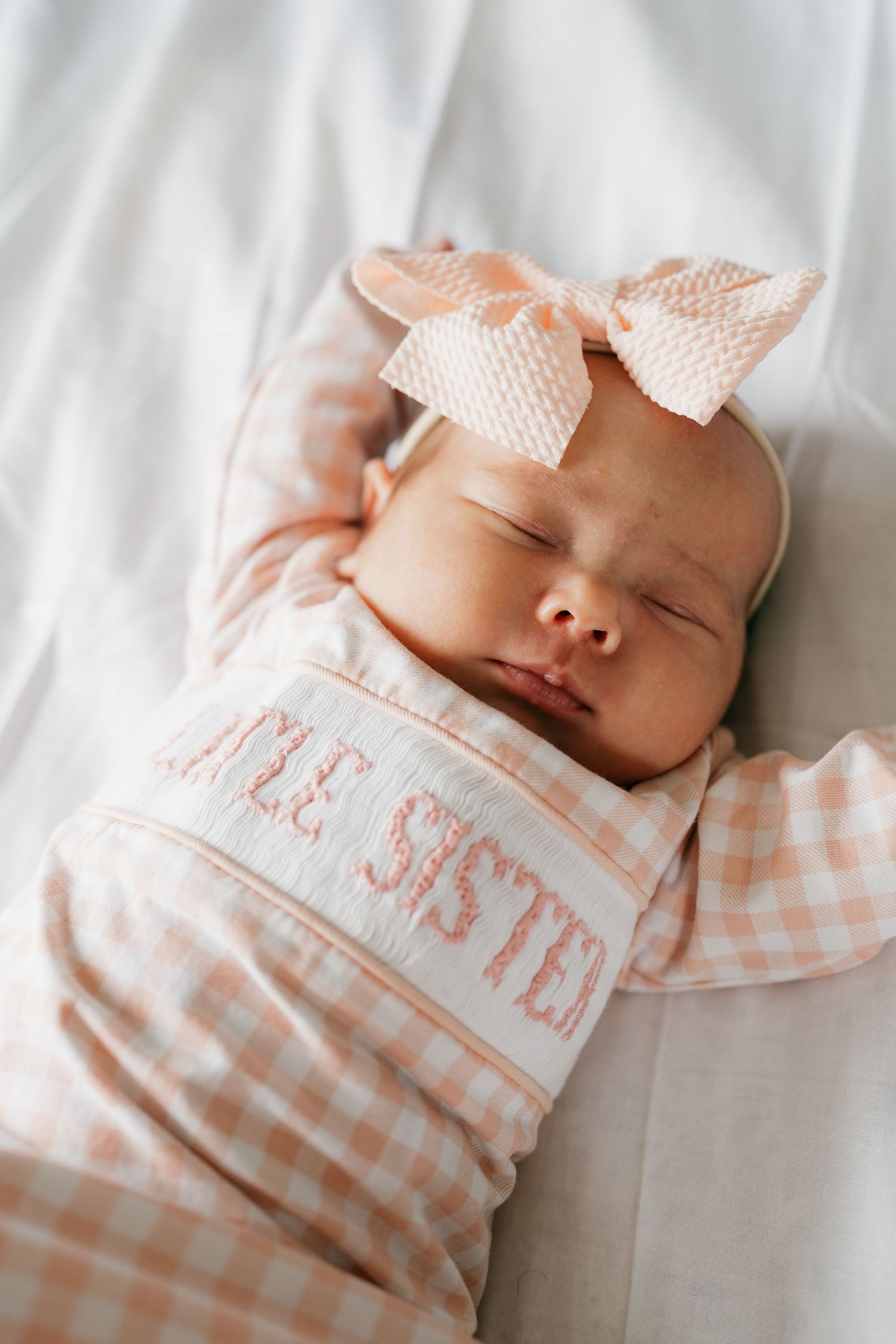 Sleeping newborn wearing a pink gingham outfit with a white bow headband on white bedding.