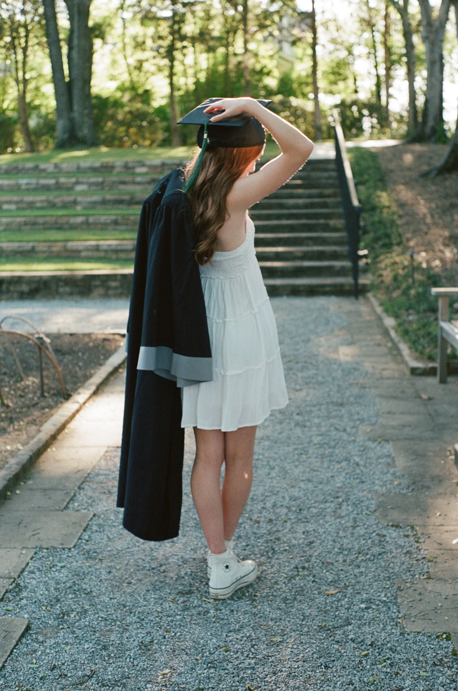 Graduate holding cap and gown while standing on stone steps in an outdoor garden setting.