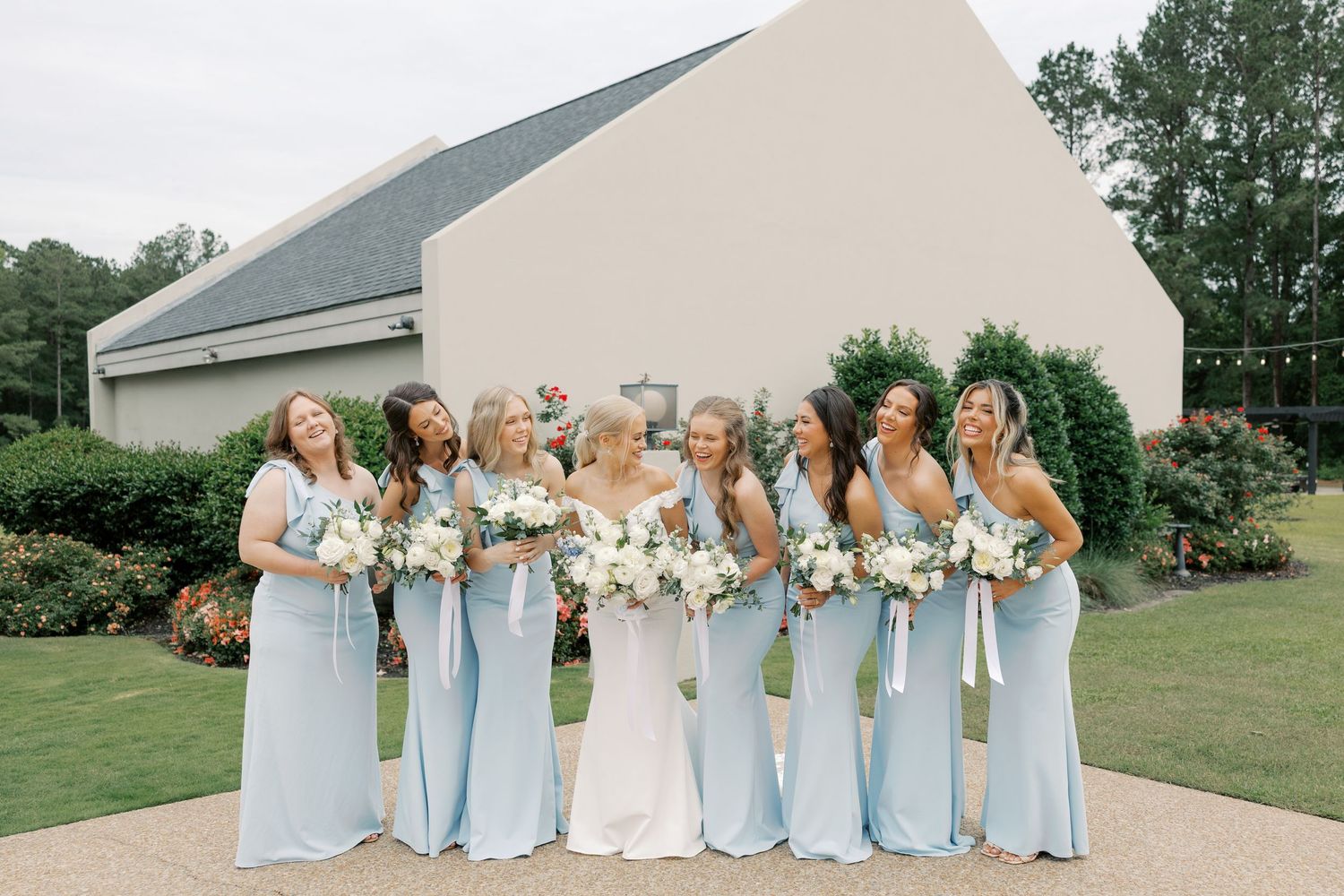 Wedding party in light blue dresses poses together outside modern white building.