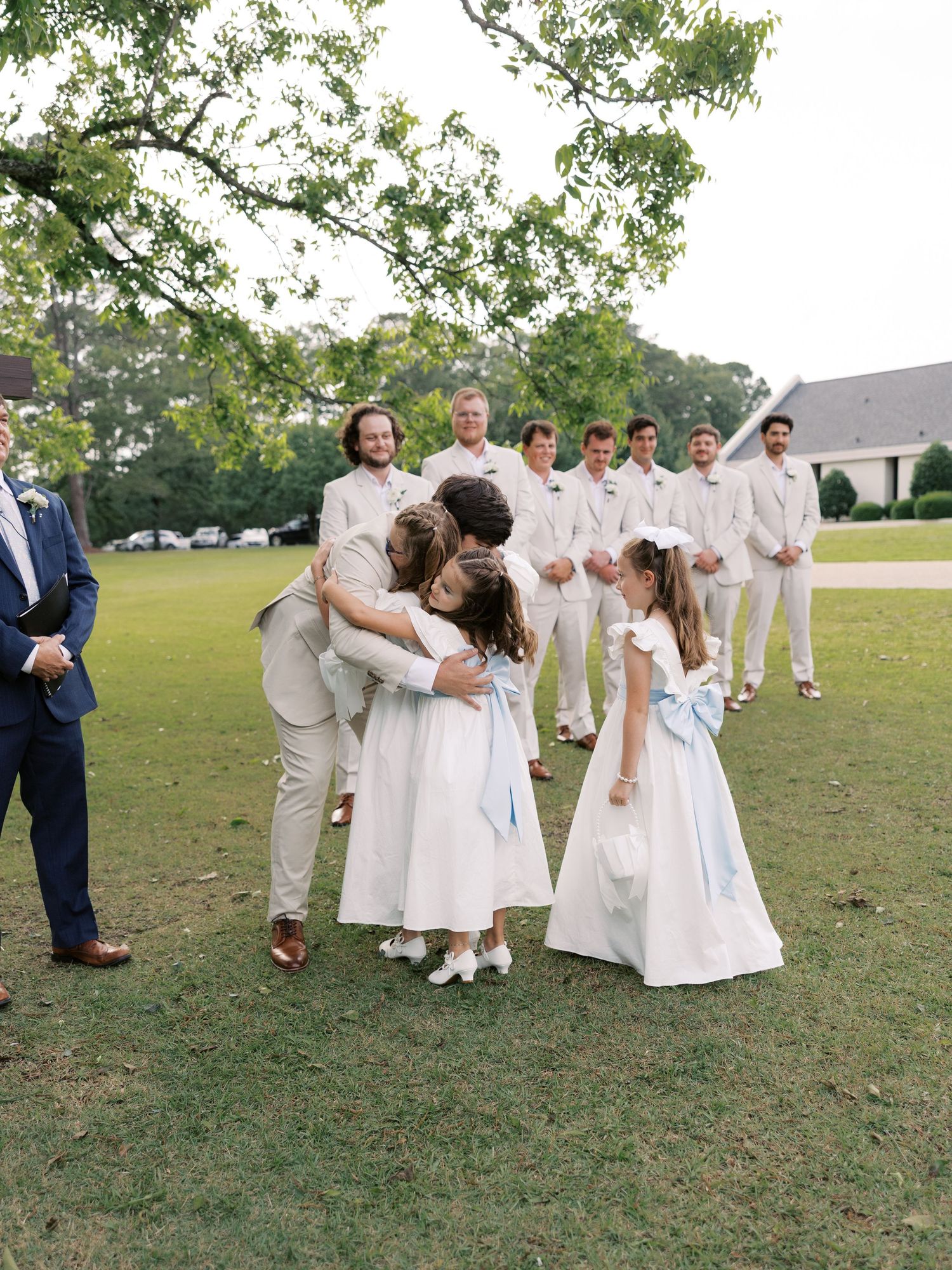 Wedding party members in white attire gather on a grassy lawn.