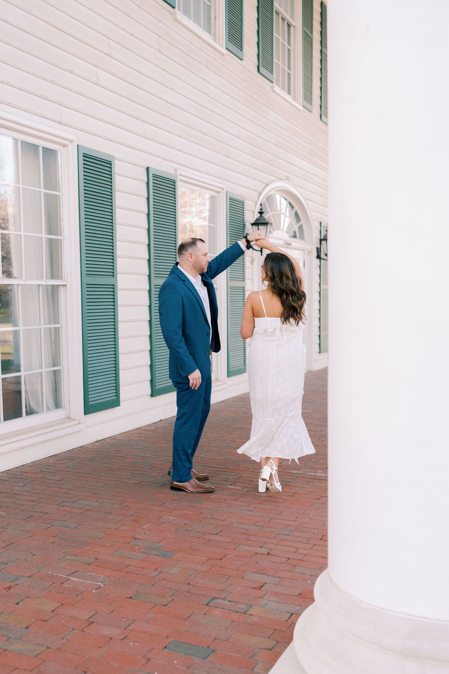Playful couple dances along white colonial building with green shutters and brick walkway.