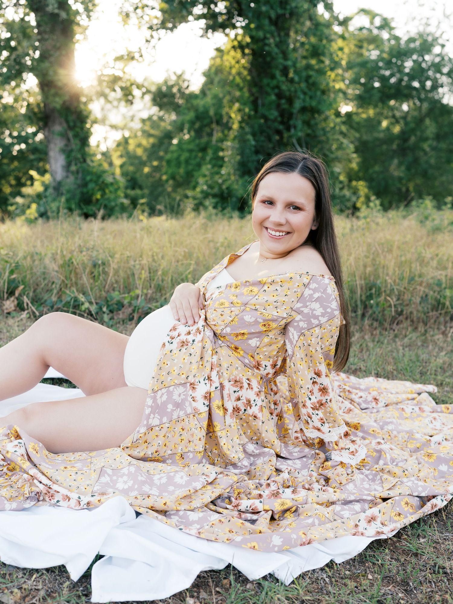 Expectant mother in yellow floral dress reclining on white blanket in sunlit field.