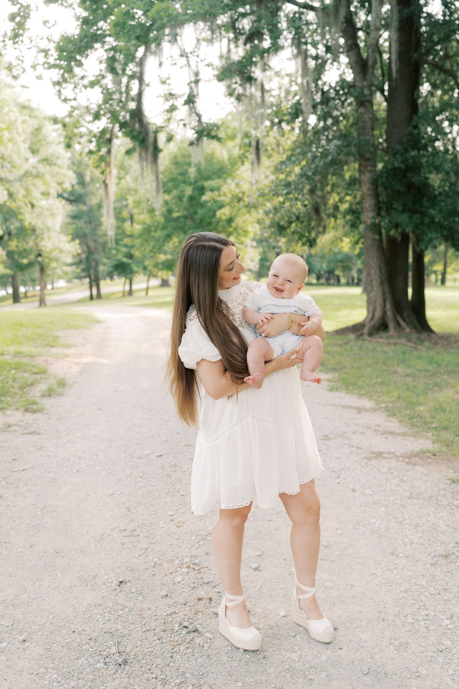 A woman in white holds a baby while standing on a sunlit forest path.