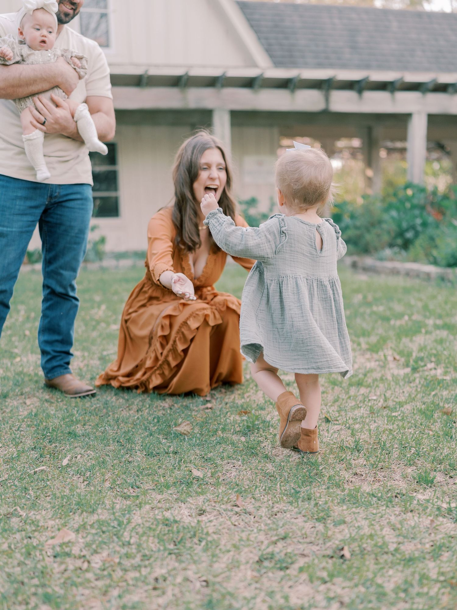 A toddler in a gray dress and brown boots walks on grass near a rustic building.