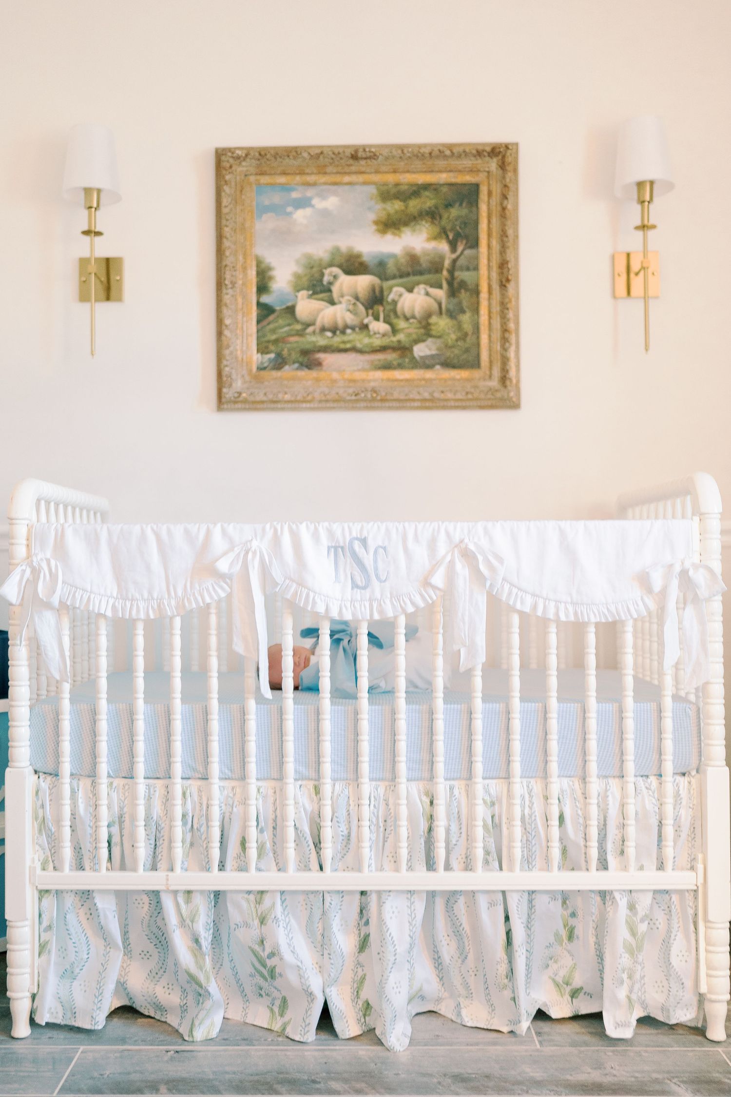 White crib bedding with delicate bows adorns a nursery with framed artwork and wall sconces.
