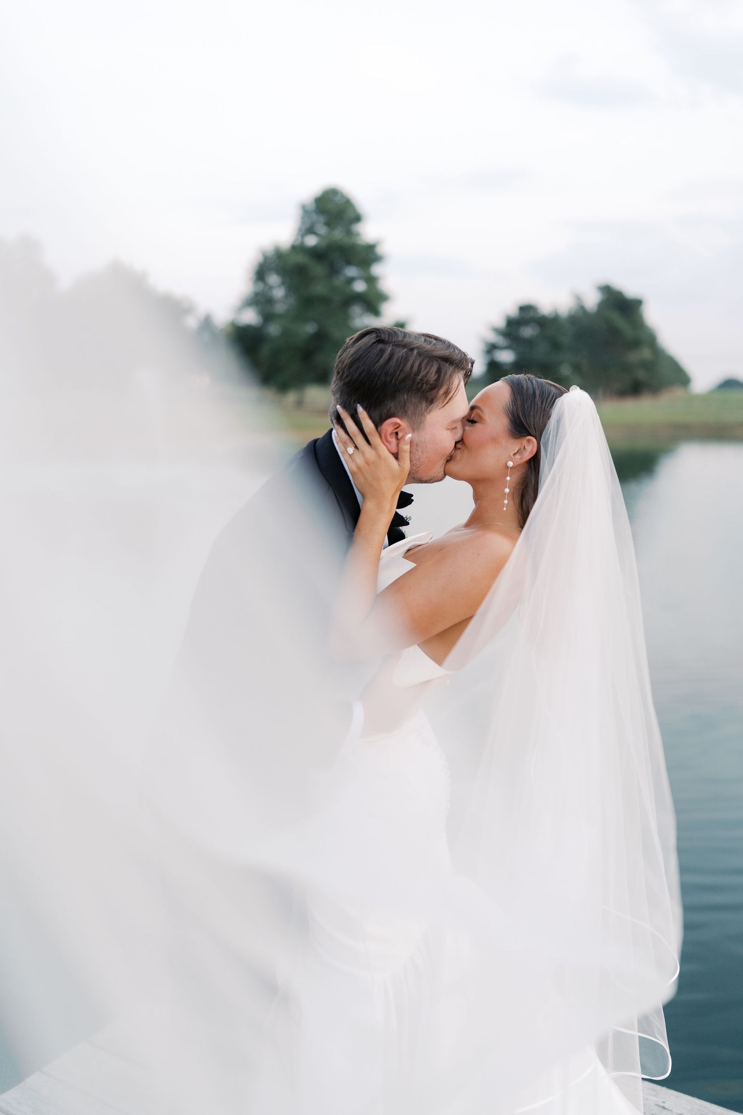 Bride and groom share an intimate moment under a flowing veil by the waterfront.