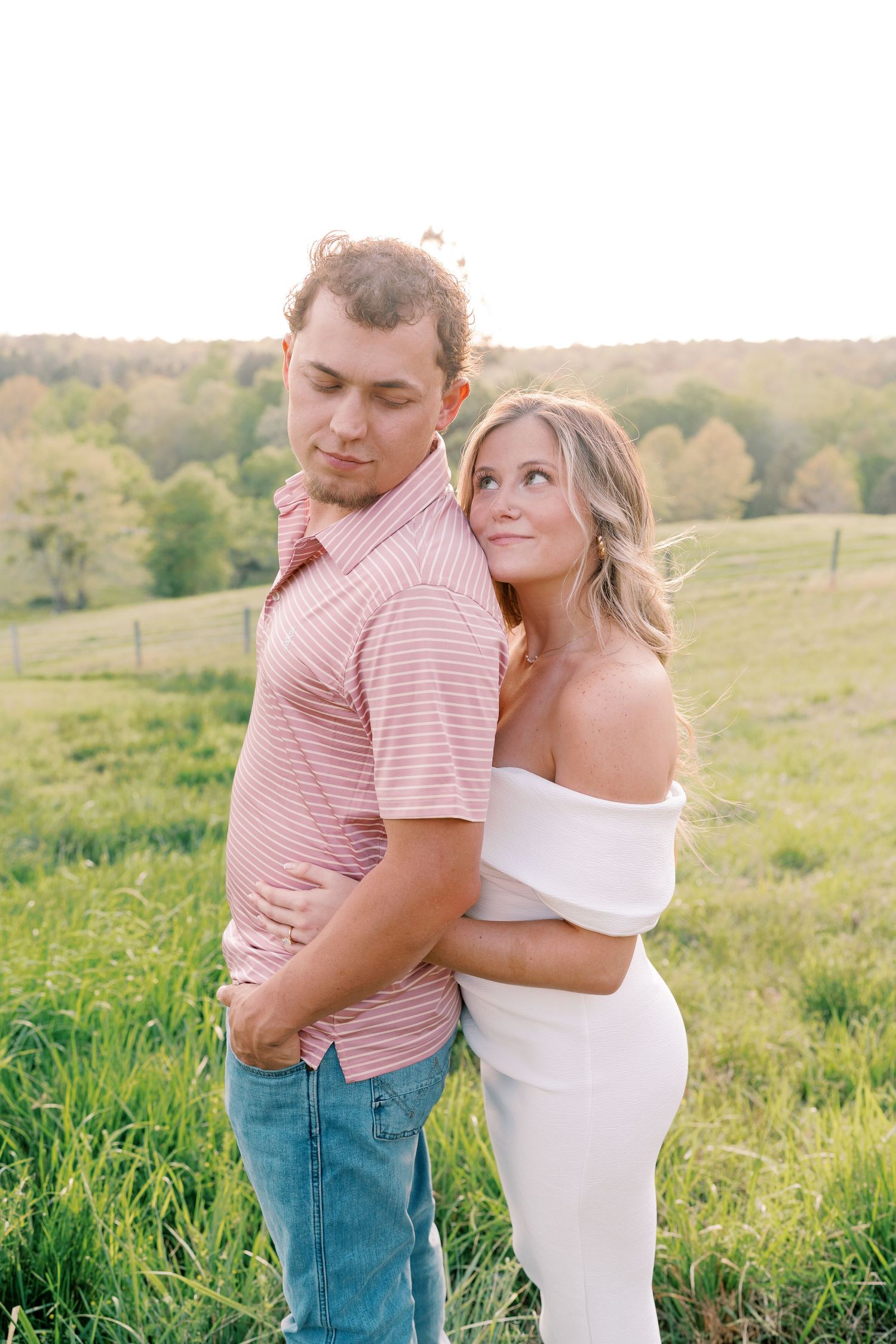 A romantic couple embraces in a sunlit meadow wearing pink and white outfits.