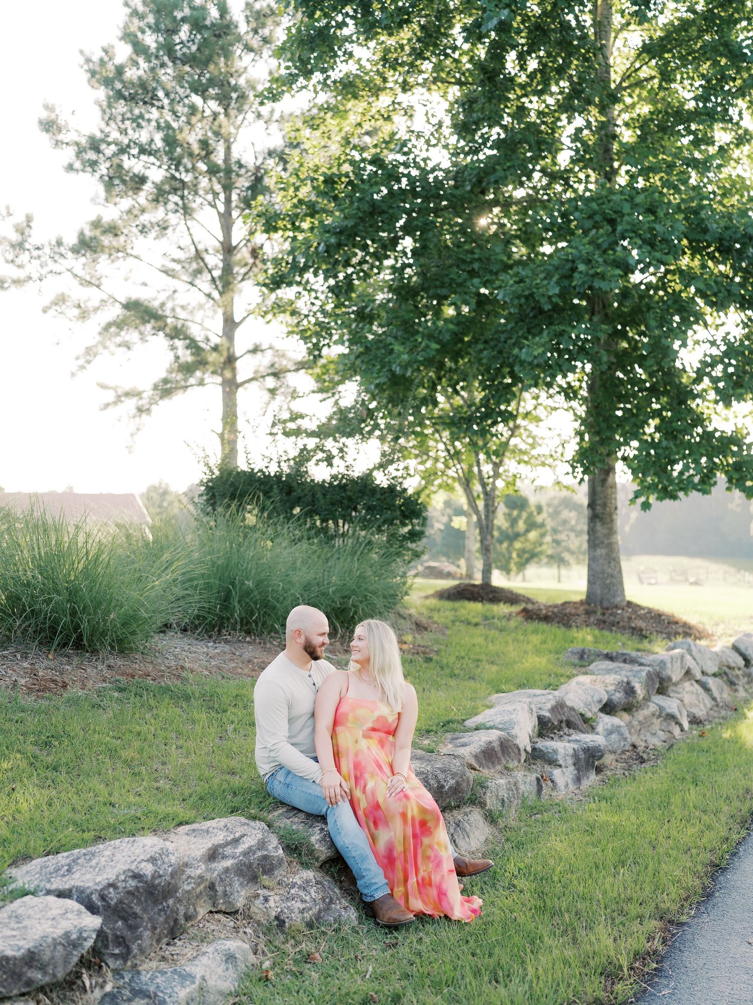 Couple relaxing on stone wall amid lush greenery and afternoon sunlight.
