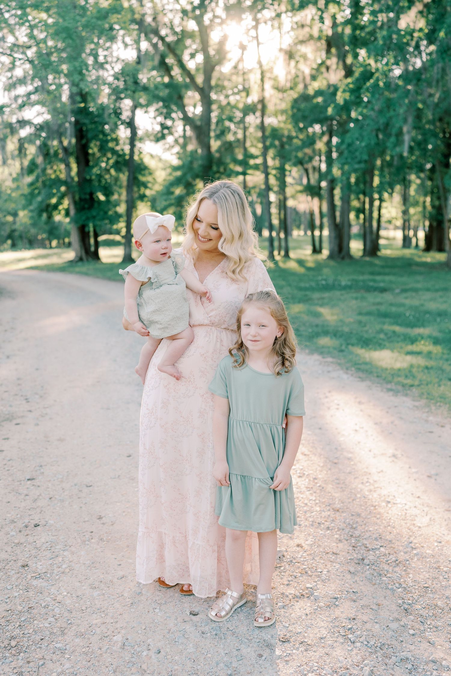 Family portrait on a sunlit path with coordinated pastel outfits in a park setting.