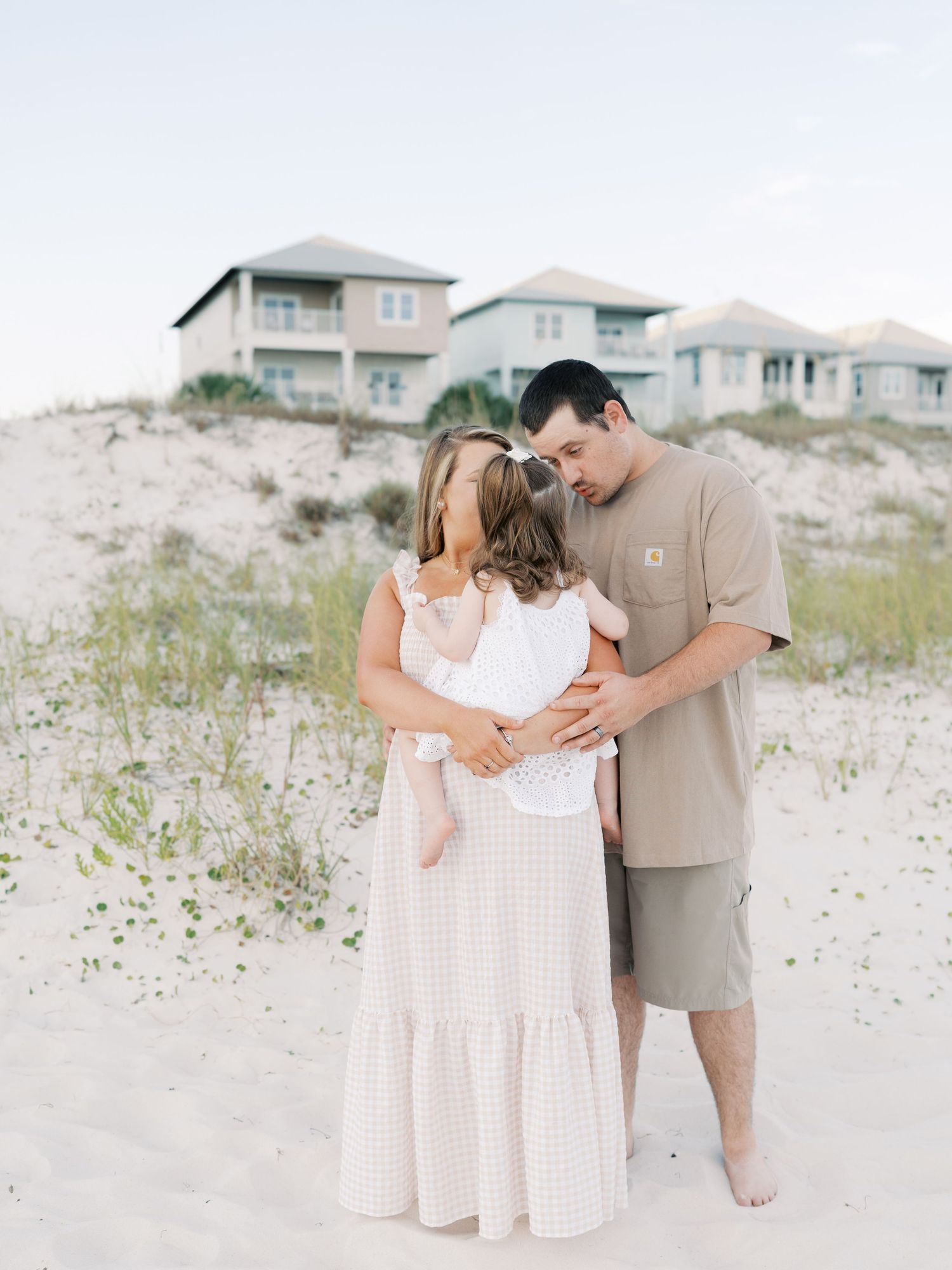Casual beach portrait session with family members embracing on sandy dunes near beach houses.