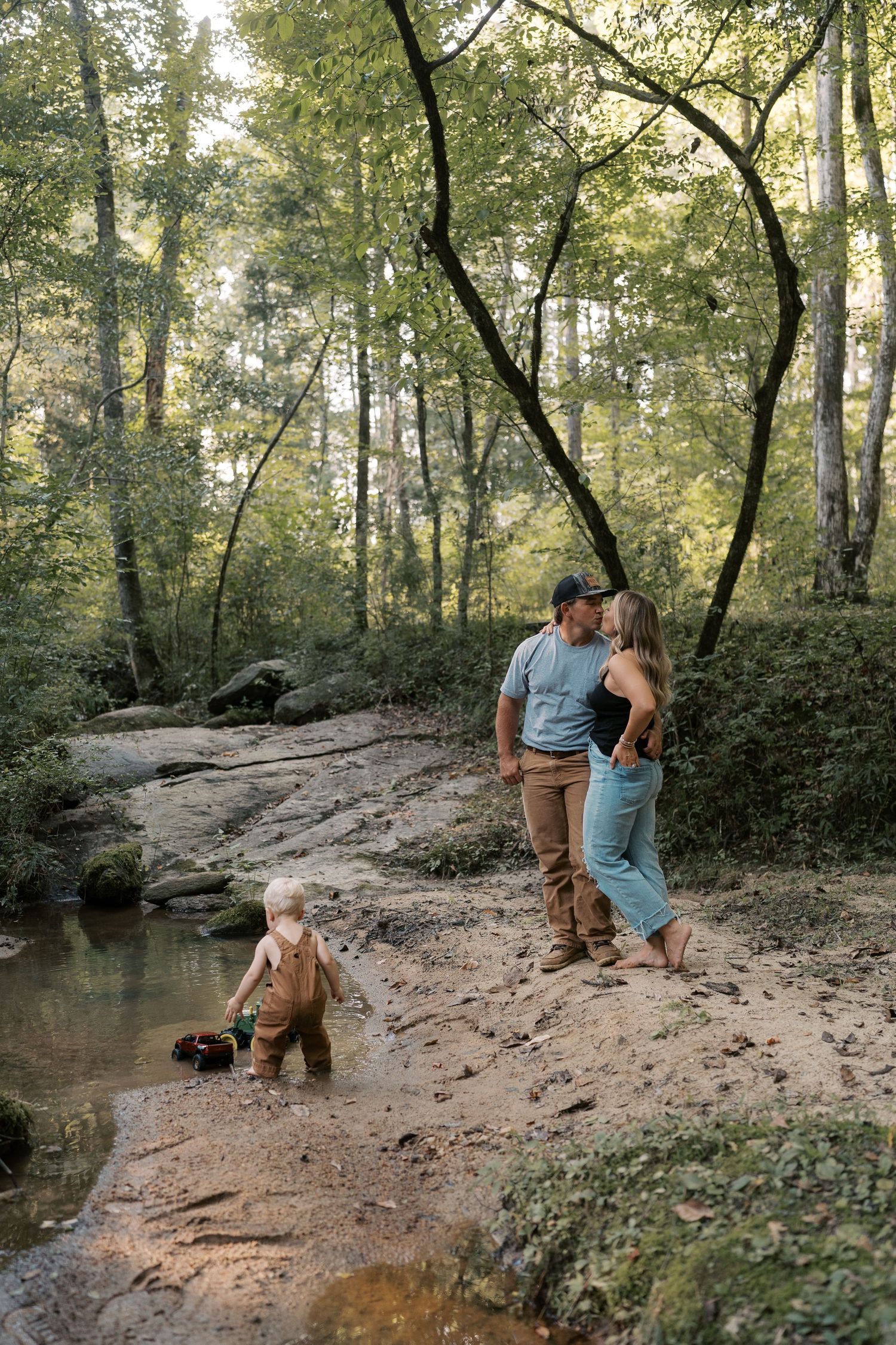 A family plays together by a creek in a wooded area on a sunny afternoon.