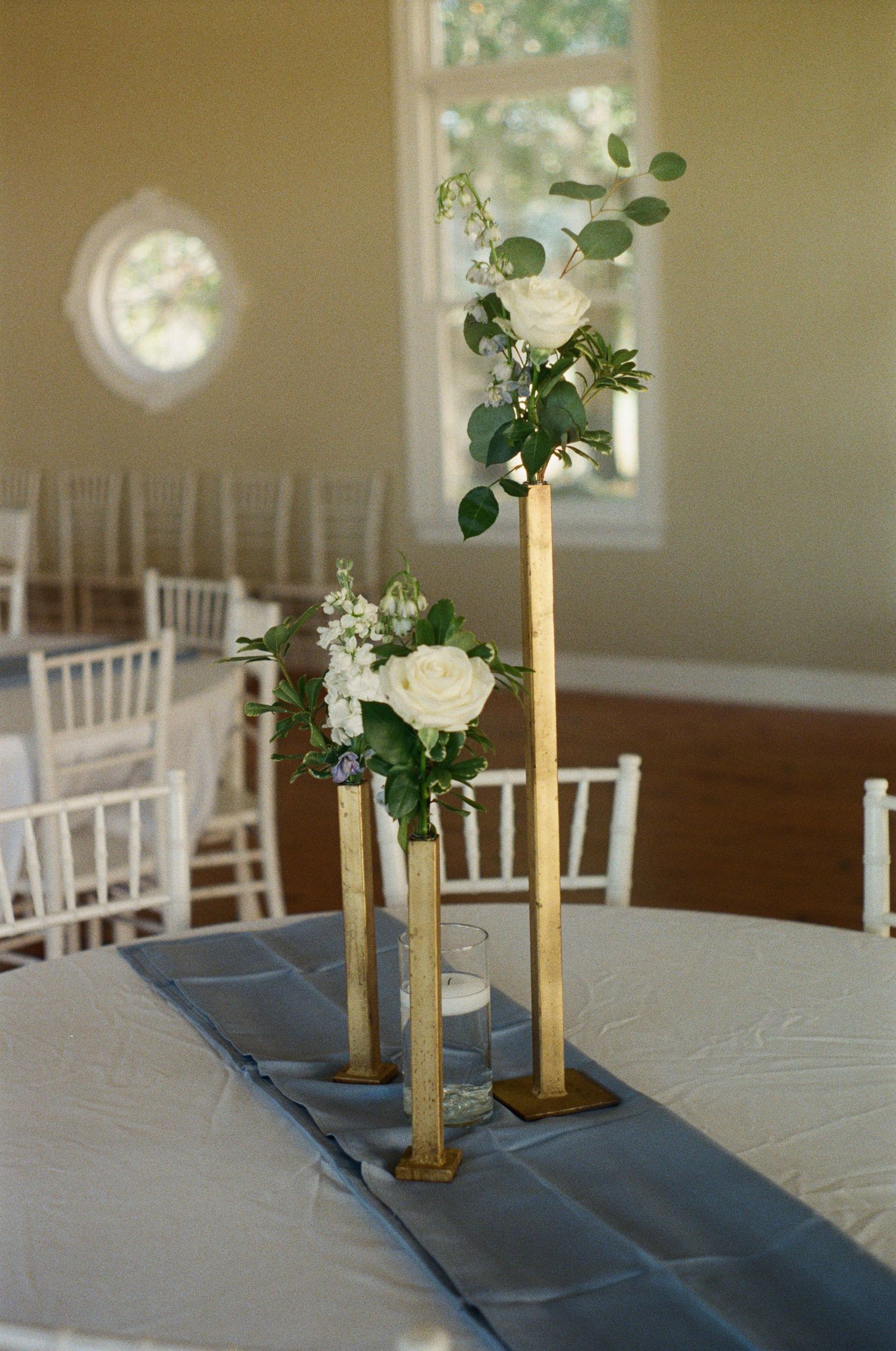 Wedding reception table with blue runner and gold vases holding white floral arrangements.