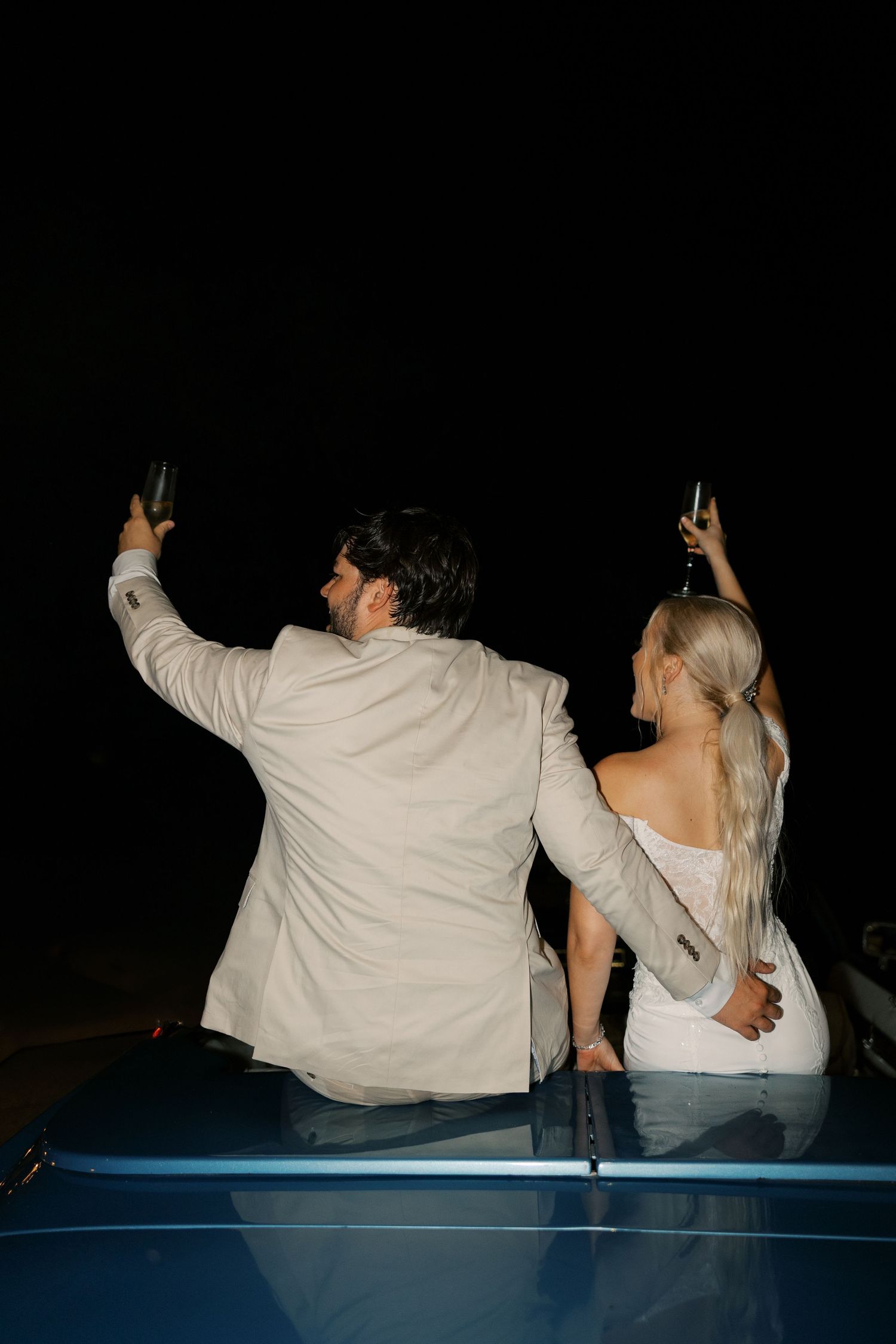Couple sits in vintage car at night with arms raised in celebration.