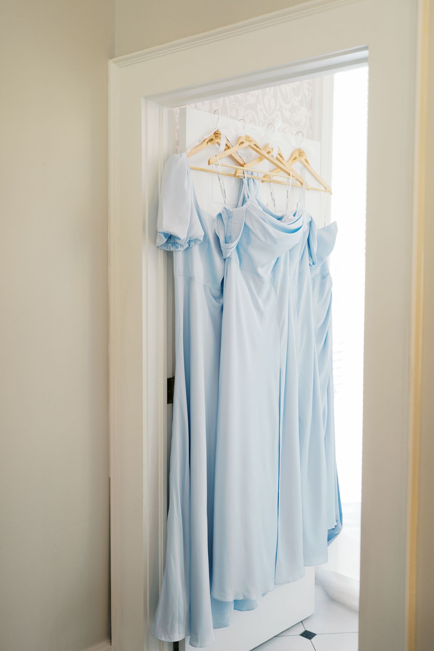 Light blue bridesmaid dresses hanging on golden hangers against a white wall.